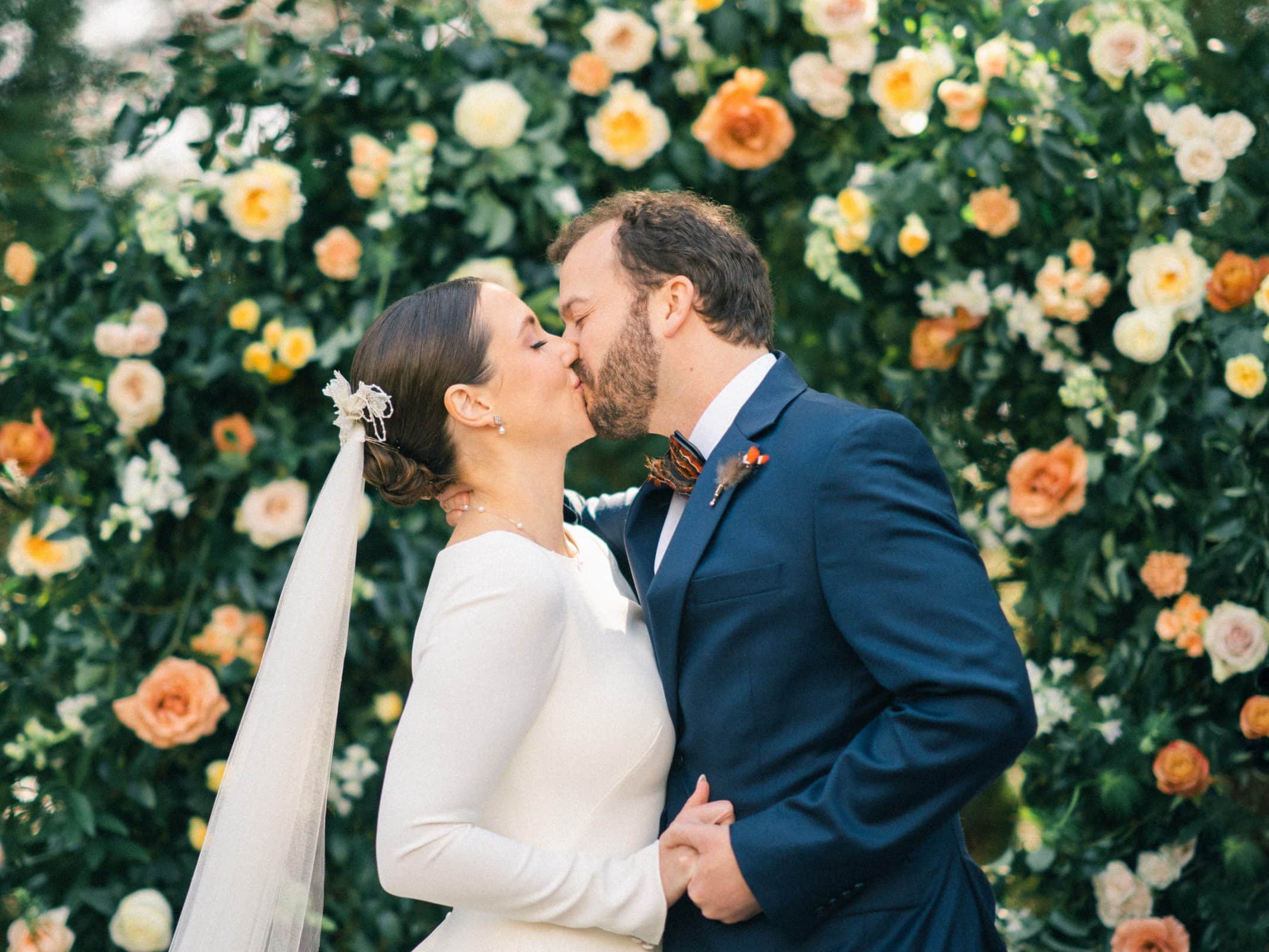 sutherland estate wedding - stephanie & hunter At their Sutherland Estate wedding, a couple in wedding attire kisses in front of a lush floral backdrop adorned with pink, white, and peach blooms. The bride's long veil flows gracefully as the groom stands by her side in a navy suit.