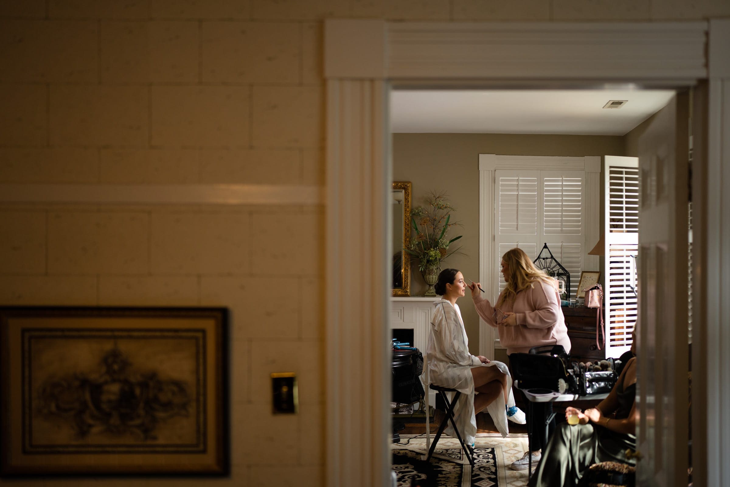 sutherland estate wedding - stephanie & hunter In a well-lit room with beige walls and cozy ambiance, a person is having their makeup done for a Sutherland Estate wedding. Sitting on chairs amid beauty equipment and decorative elements, the scene exudes elegance and anticipation for the special day.