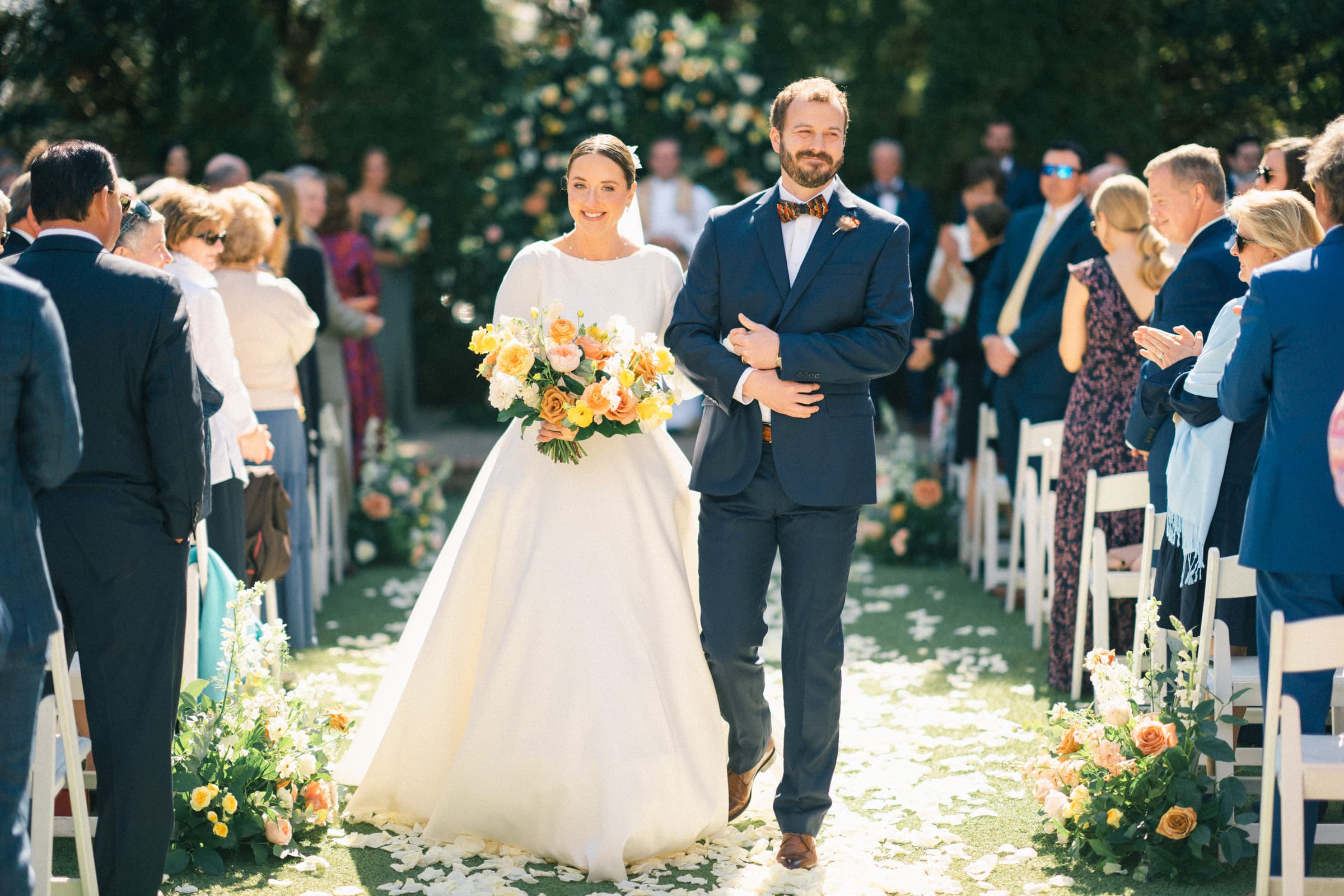 sutherland estate wedding - stephanie & hunter A bride in a white dress and a groom in a navy suit walk down the outdoor aisle at Sutherland Estate, lined with chairs and flower petals. Surrounded by standing guests, the bride holds a bouquet of yellow and orange flowers as sunlight filters through the trees.
