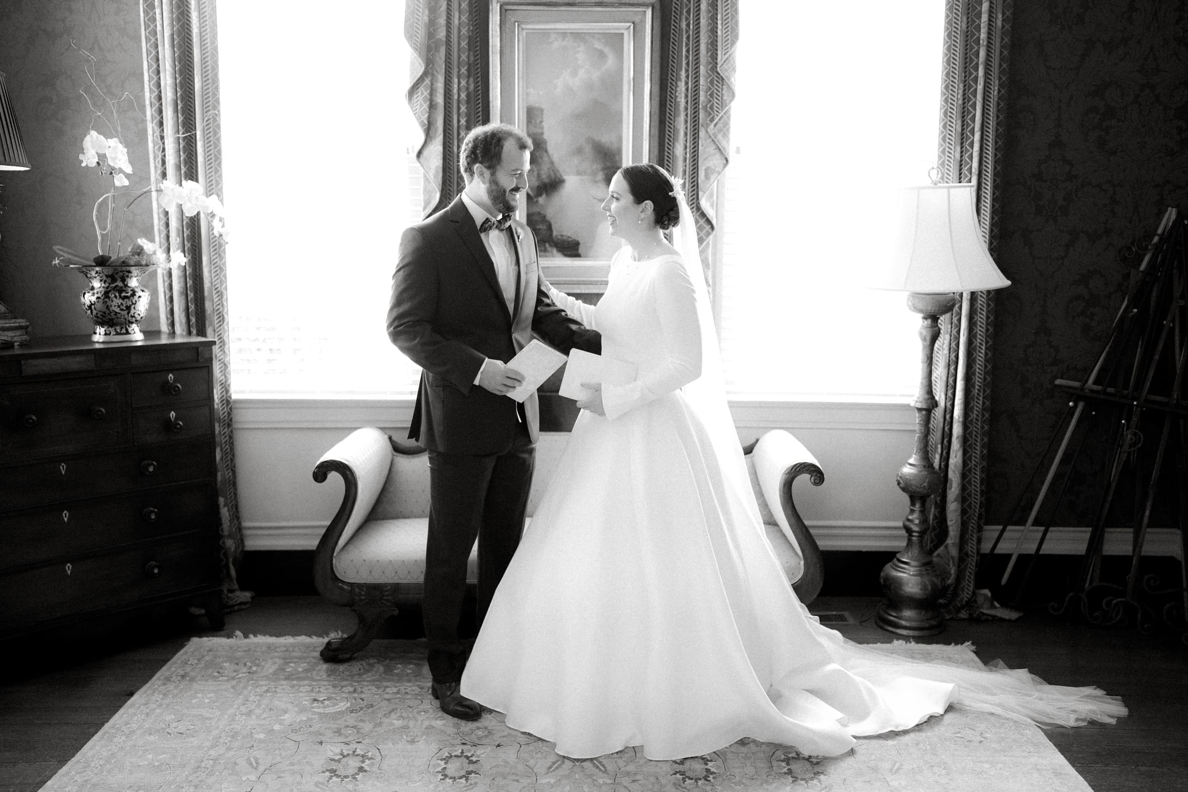 sutherland estate wedding - stephanie & hunter A bride and groom stand in a room at Sutherland Estate, exchanging letters and smiling warmly. The vintage sofa, potted flowers, and elegant curtains create an intimate and classic atmosphere that perfectly encapsulates their special day.