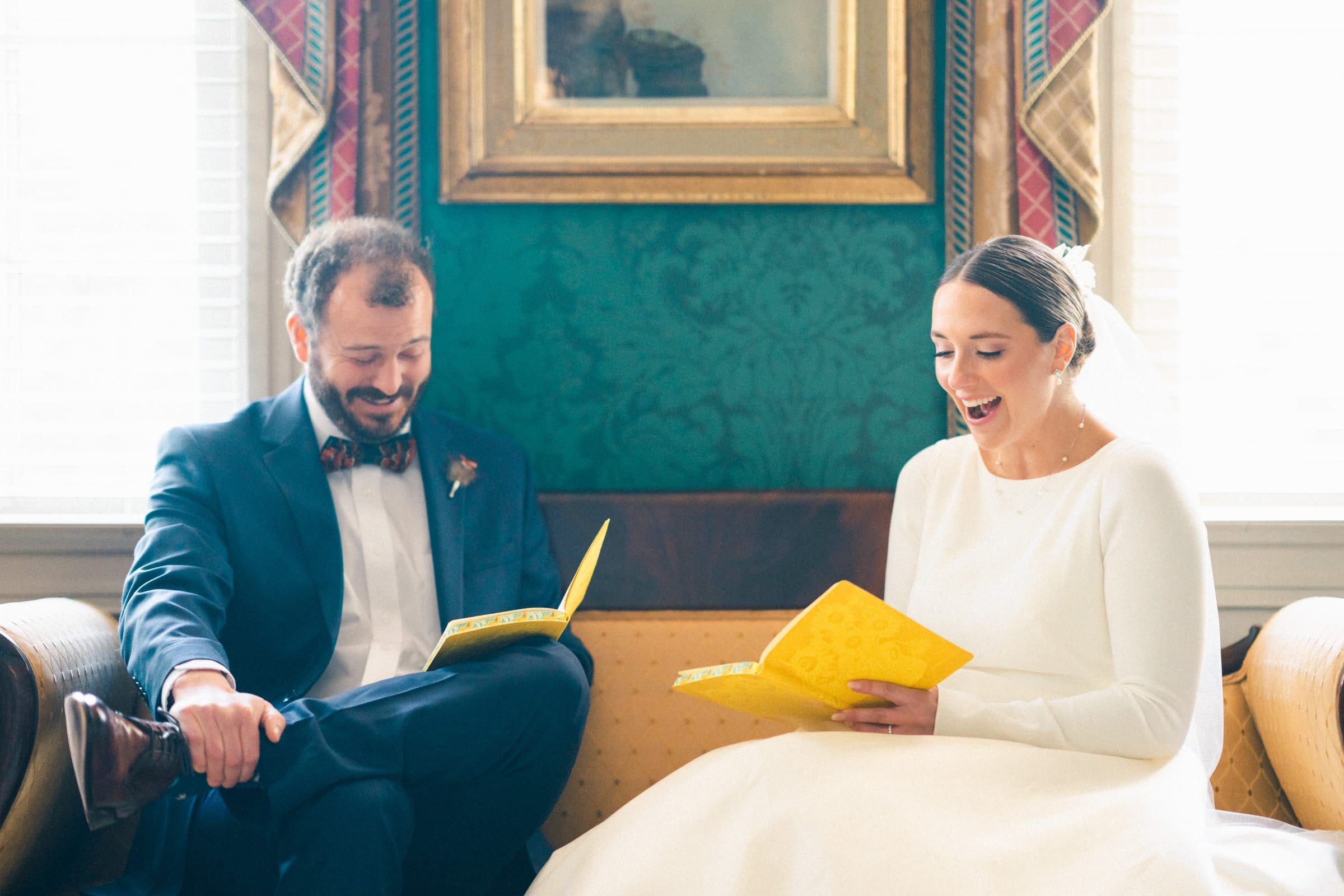 sutherland estate wedding - stephanie & hunter A bride and groom sit on a couch at their Sutherland Estate wedding, each holding a yellow booklet. The bride, in a white dress, smiles warmly, while the groom, in a navy suit, looks at his booklet with a cheerful expression. A framed painting hangs on the green wall behind them.