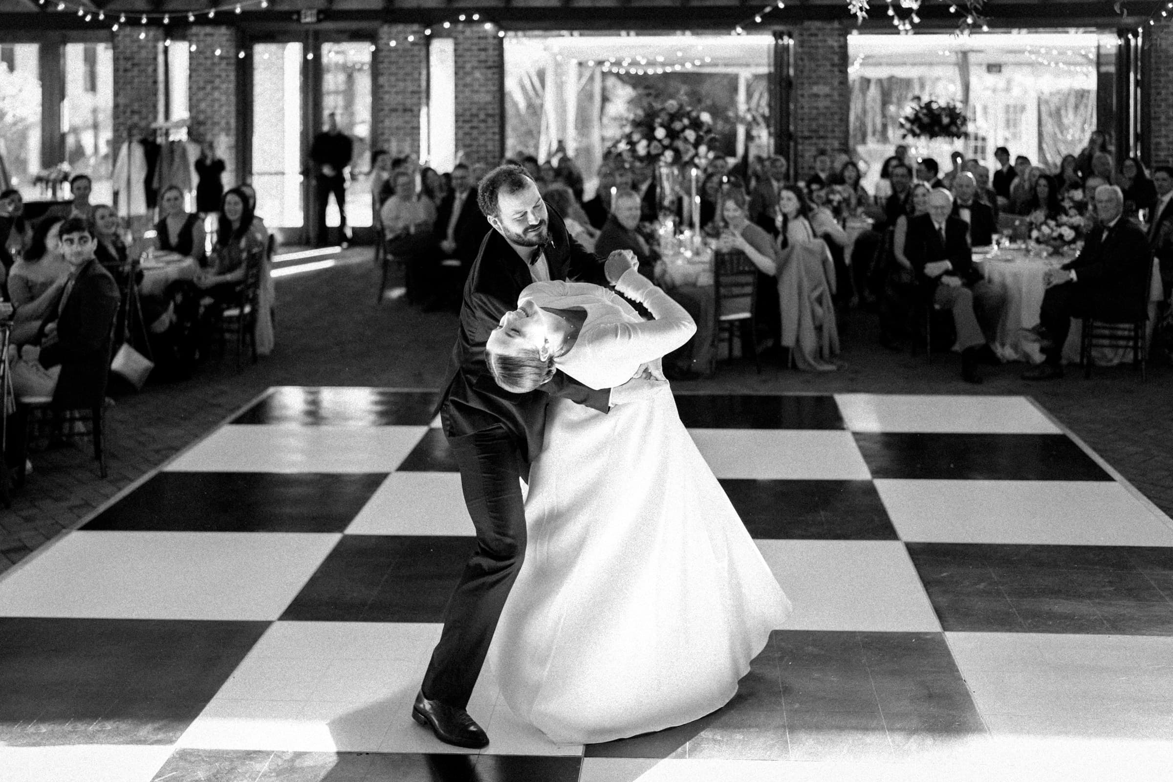 sutherland estate wedding - stephanie & hunter A couple in wedding attire is dancing on a black-and-white checkered floor at their Sutherland Estate wedding. The groom dips the bride elegantly as guests seated around the dance floor watch and smile, while string lights cast a warm glow over the enchanting scene.