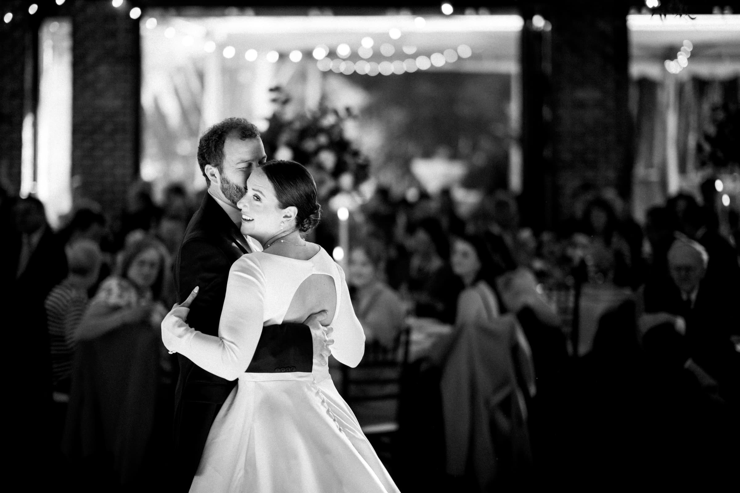 sutherland estate wedding - stephanie & hunter At a Sutherland Estate wedding, a bride and groom share a dance in a warmly lit venue. The bride dazzles in an open-back gown, while the groom looks sharp in his suit. Guests sit captivated, watching the couple under string lights that add to the festive atmosphere.