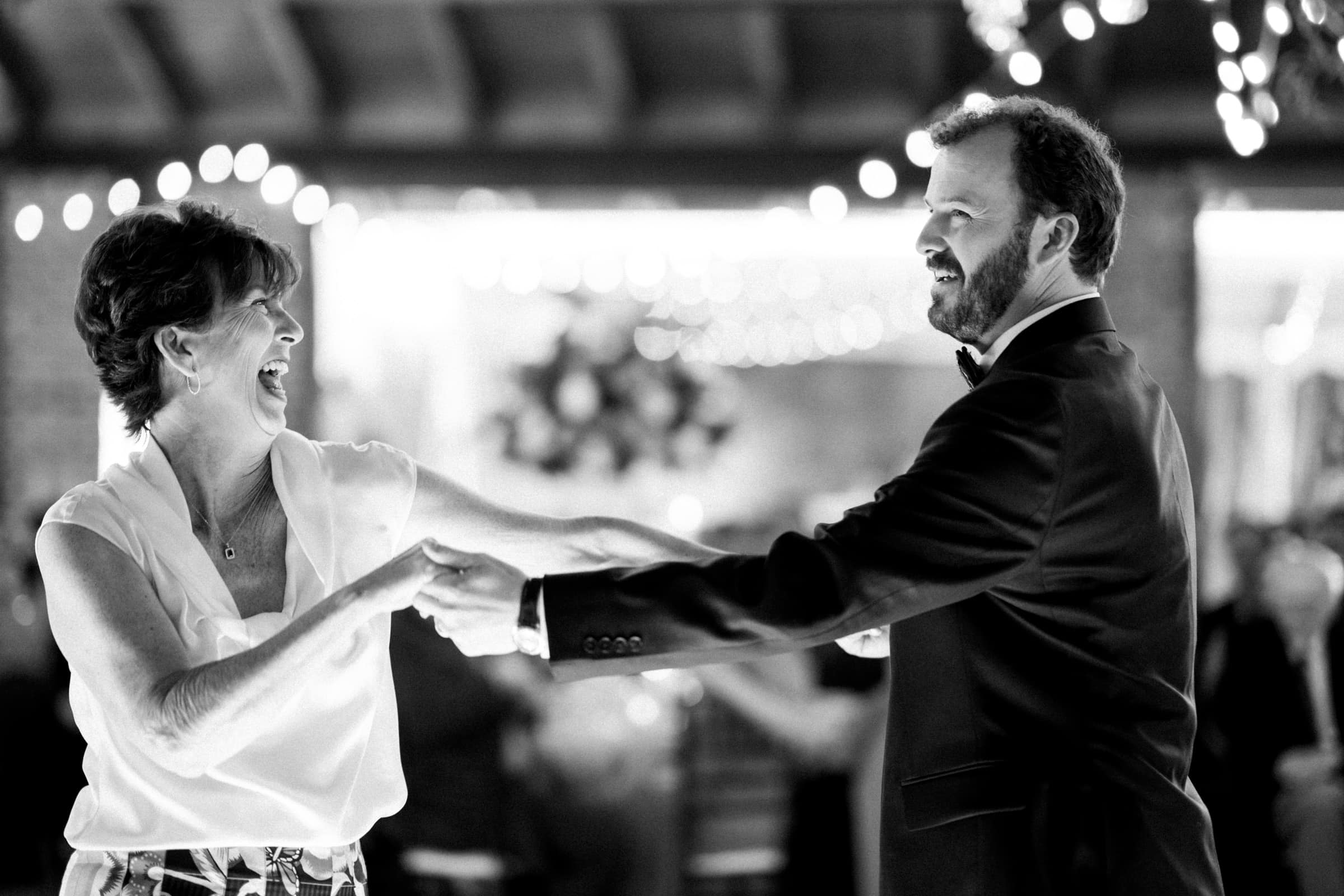 sutherland estate wedding - stephanie & hunter A woman and a man dance joyfully in a warmly lit venue at a Sutherland Estate wedding. The woman is wearing a sleeveless blouse, and the man is in a suit. They smile at each other, surrounded by blurred lights and people in the background. The image is in black and white.