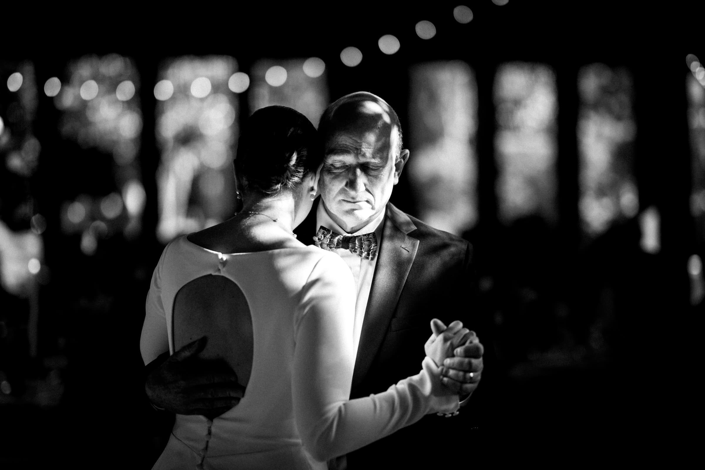 sutherland estate wedding - stephanie & hunter A black and white photo captures a couple dancing closely at a Sutherland Estate wedding. The woman, in an open-back dress, shares an intimate moment with the man in a suit. Soft, blurred lights twinkle gently in the background.