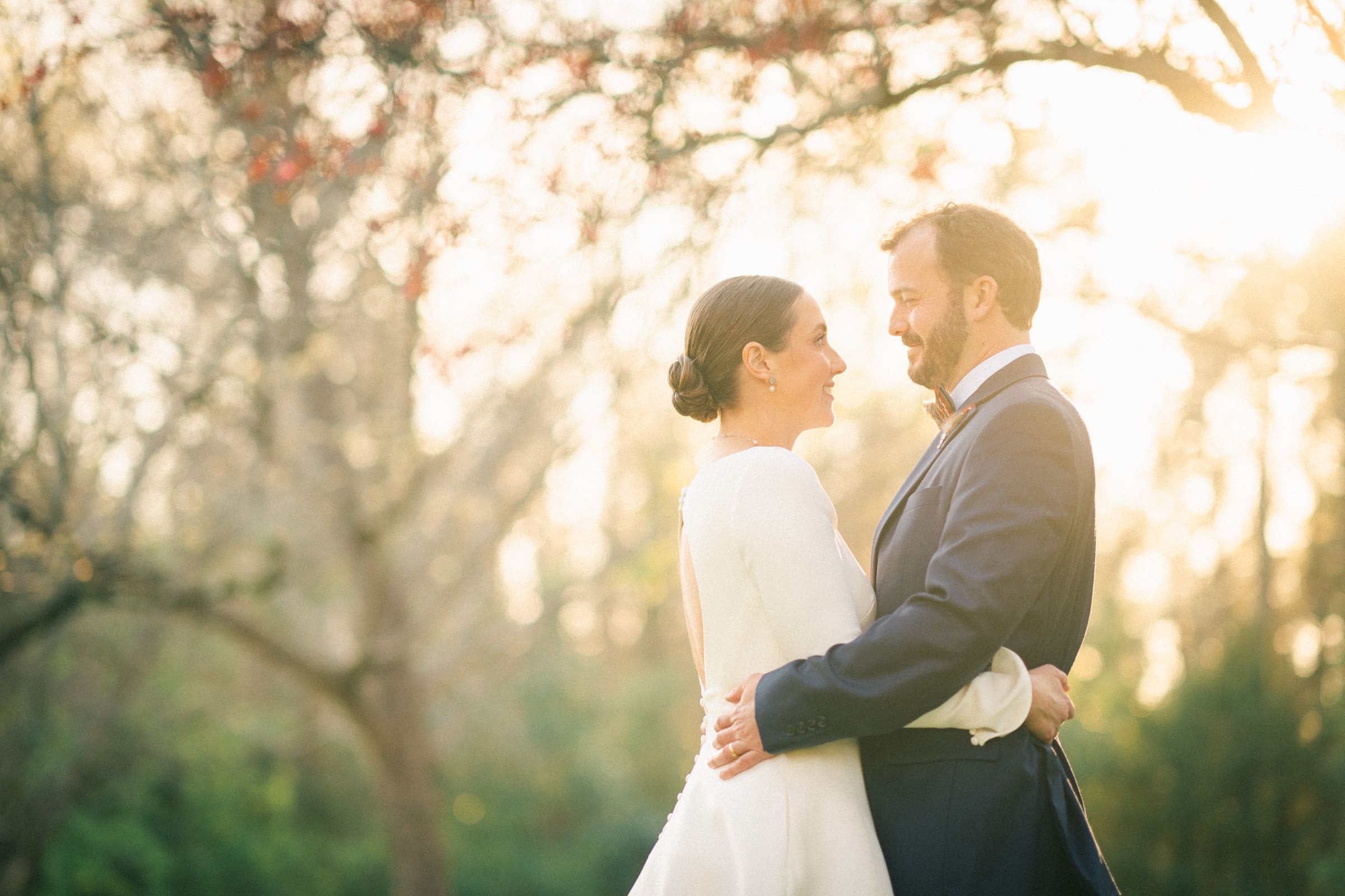 sutherland estate wedding - stephanie & hunter Amidst the soft, golden lighting and blurred trees of a Sutherland Estate wedding, a couple in formal attire embraces outdoors. The woman's white dress and the man's dark suit complement their graceful smiles as they share an intimate moment.