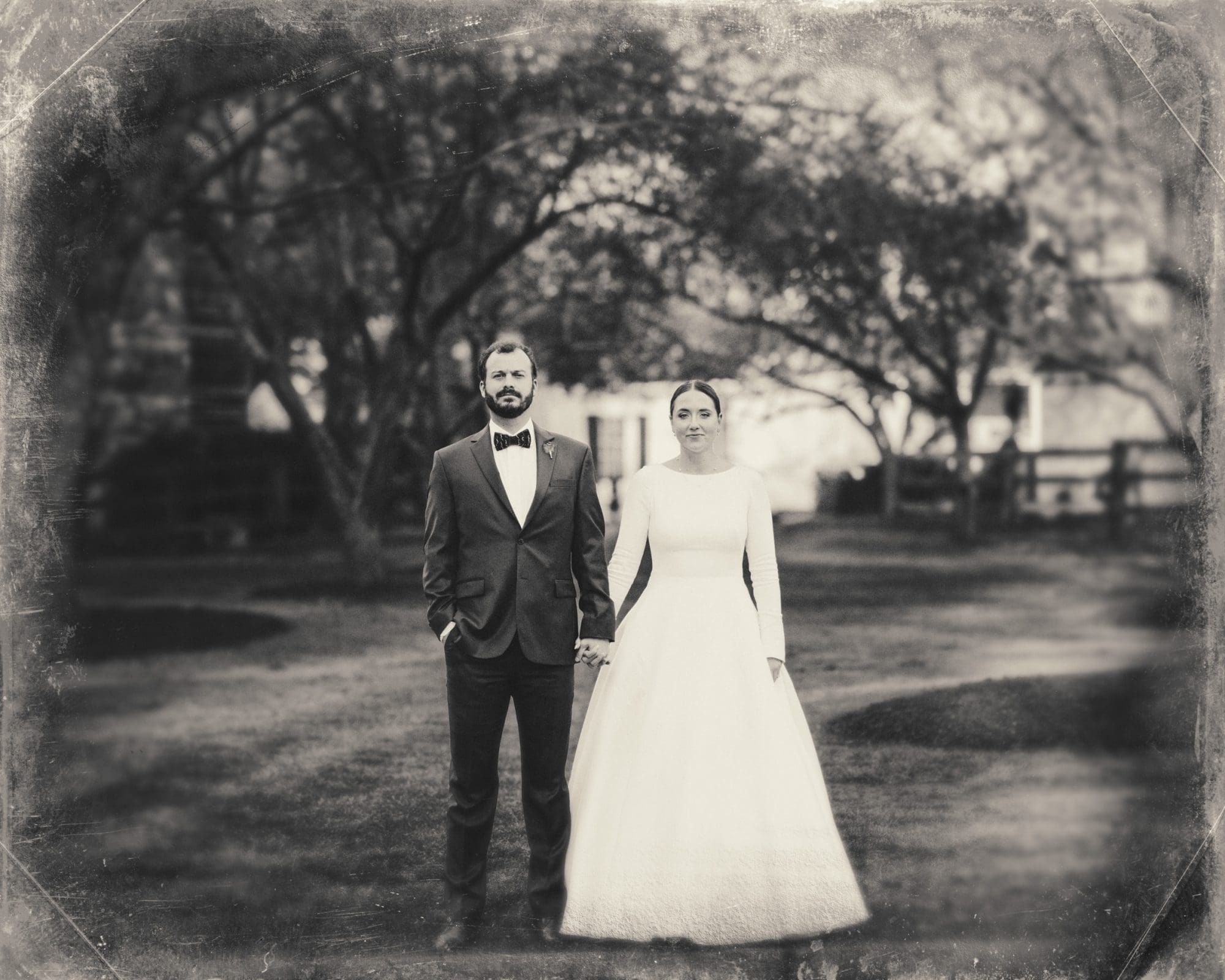 sutherland estate wedding - stephanie & hunter A vintage-style photograph captures a newlywed couple at their Sutherland Estate wedding, standing outdoors. The groom wears a dark suit with a bow tie, and the bride dons a long, white gown. They hold hands against a blurred, leafy backdrop.