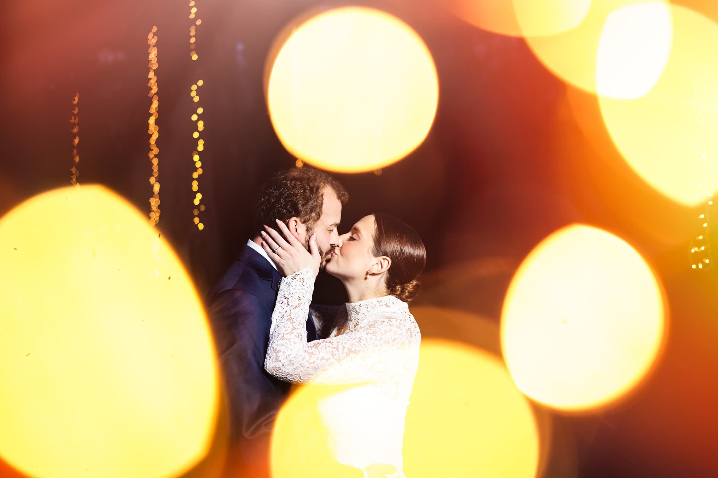 sutherland estate wedding - stephanie & hunter A couple shares a kiss at their Sutherland Estate wedding, surrounded by large, blurred golden lights that create a romantic and dreamy atmosphere. The woman is in a white lace dress, and the man is in a dark suit.