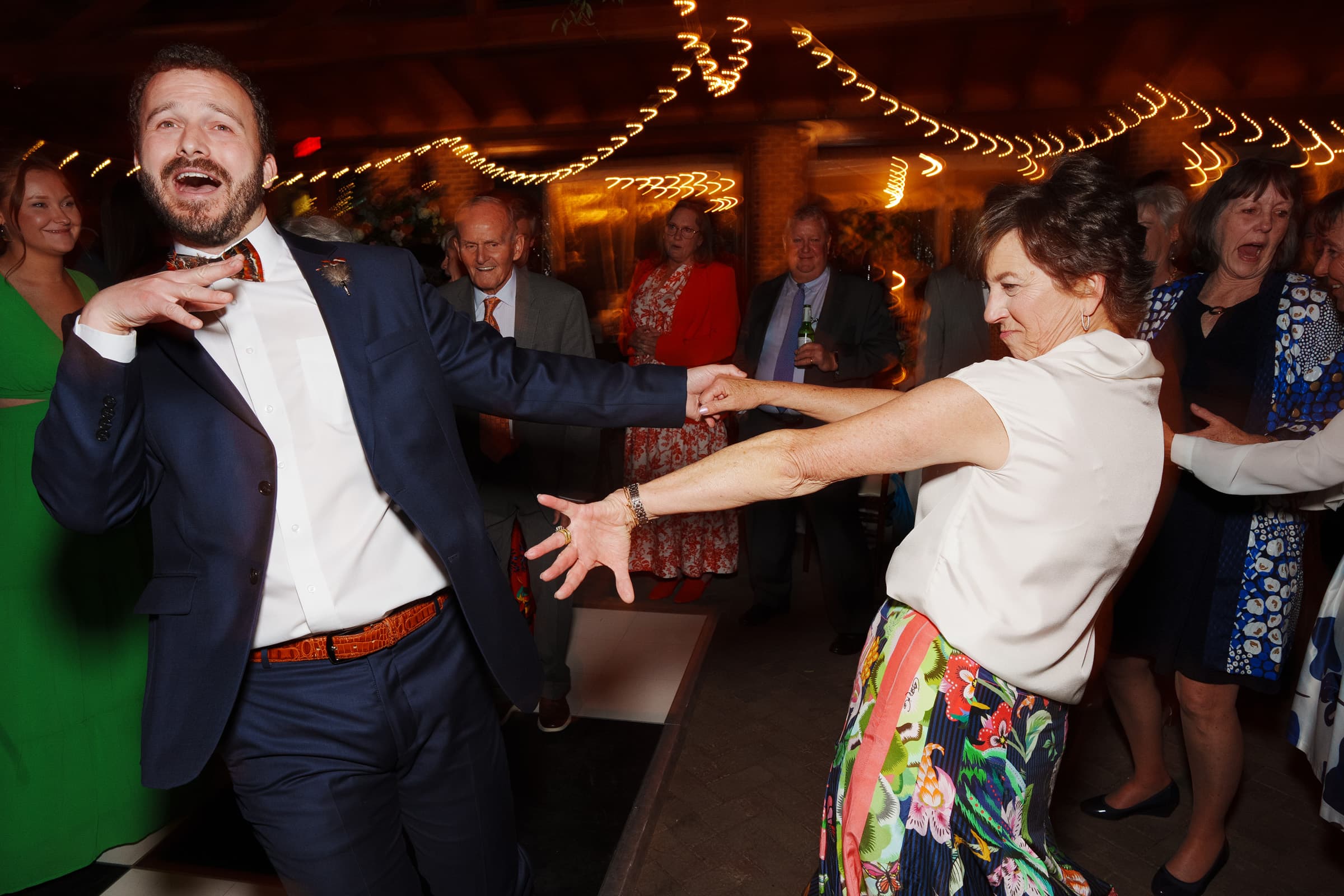 sutherland estate wedding - stephanie & hunter A joyful scene at a Sutherland Estate wedding shows a man in a suit and a woman in a colorful skirt dancing energetically. Other guests, some smiling and clapping, are visible in the background under string lights.