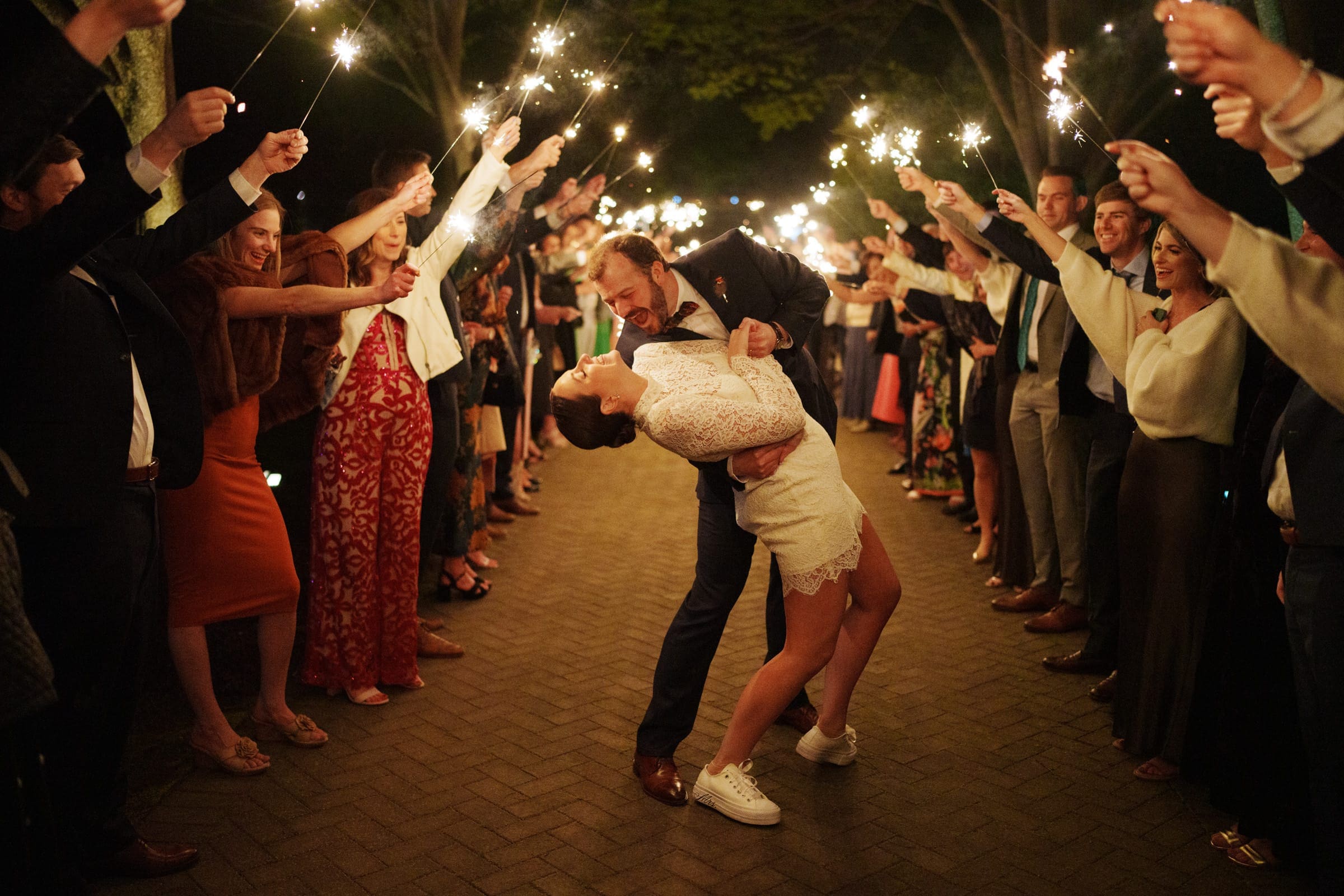 sutherland estate wedding - stephanie & hunter A bride and groom share a joyful dance on the path at Sutherland Estate, lined with friends holding sparklers. The couple's playful pose and the warm glow of the sparklers create a festive, romantic atmosphere.