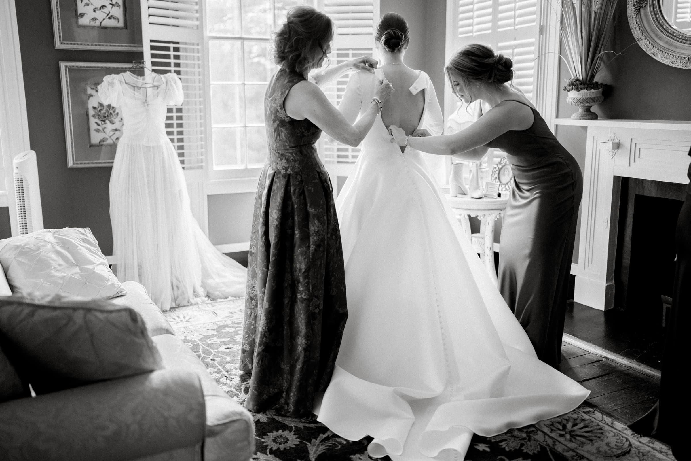 sutherland estate wedding - stephanie & hunter A bride in a long, elegant gown is being helped into her dress by two women in a cozy room at Sutherland Estate Wedding. A vintage dress is displayed nearby. The setting includes a couch, fireplace, and large windows with shutters, adding a classic and intimate atmosphere.