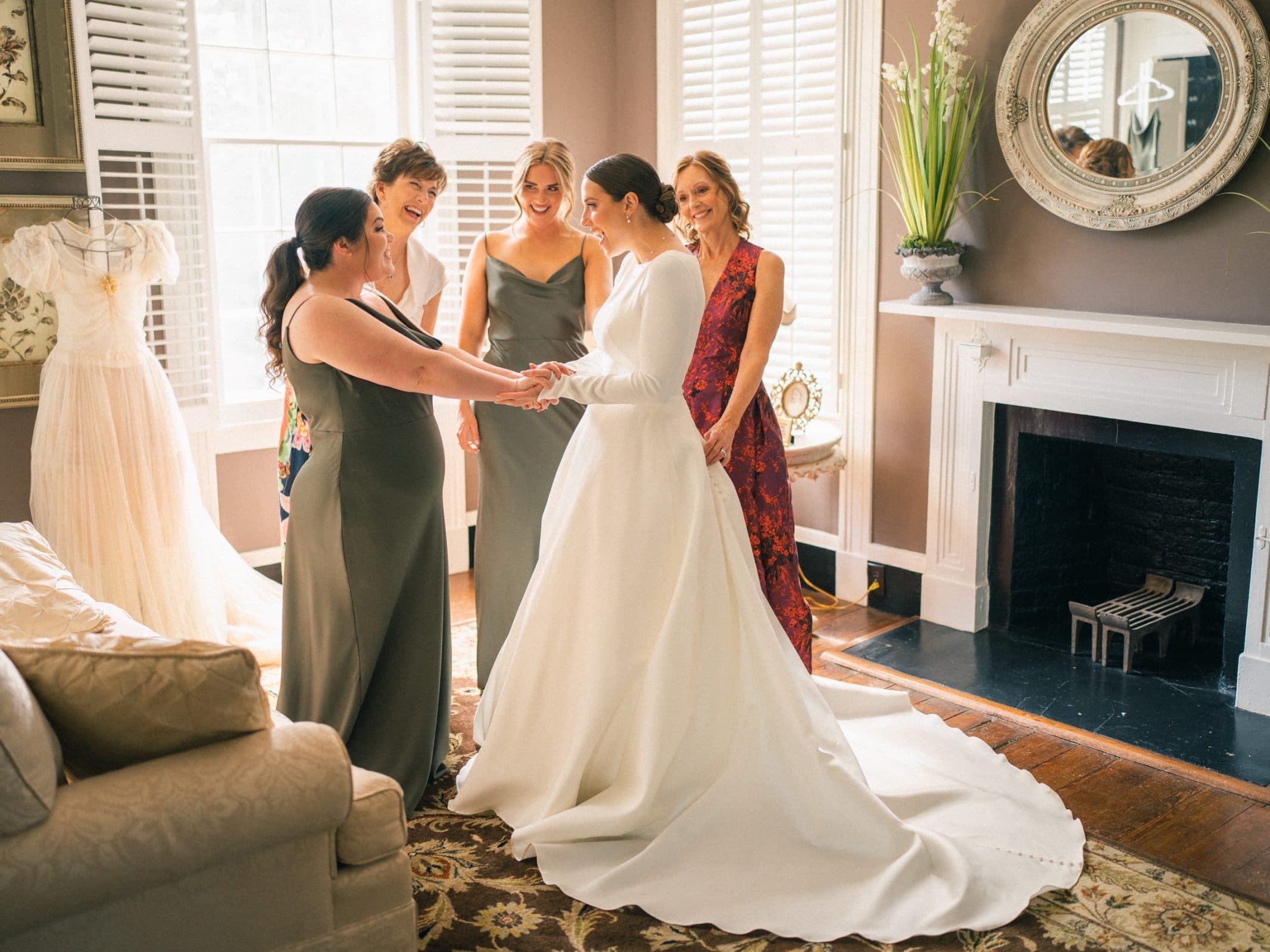 In a warmly lit room at Sutherland Estate, a bride in a white gown stands surrounded by four women, two in gray dresses and two in colorful ones. They smile as they admire her, while a mannequin adorned with a wedding dress lingers subtly in the background.