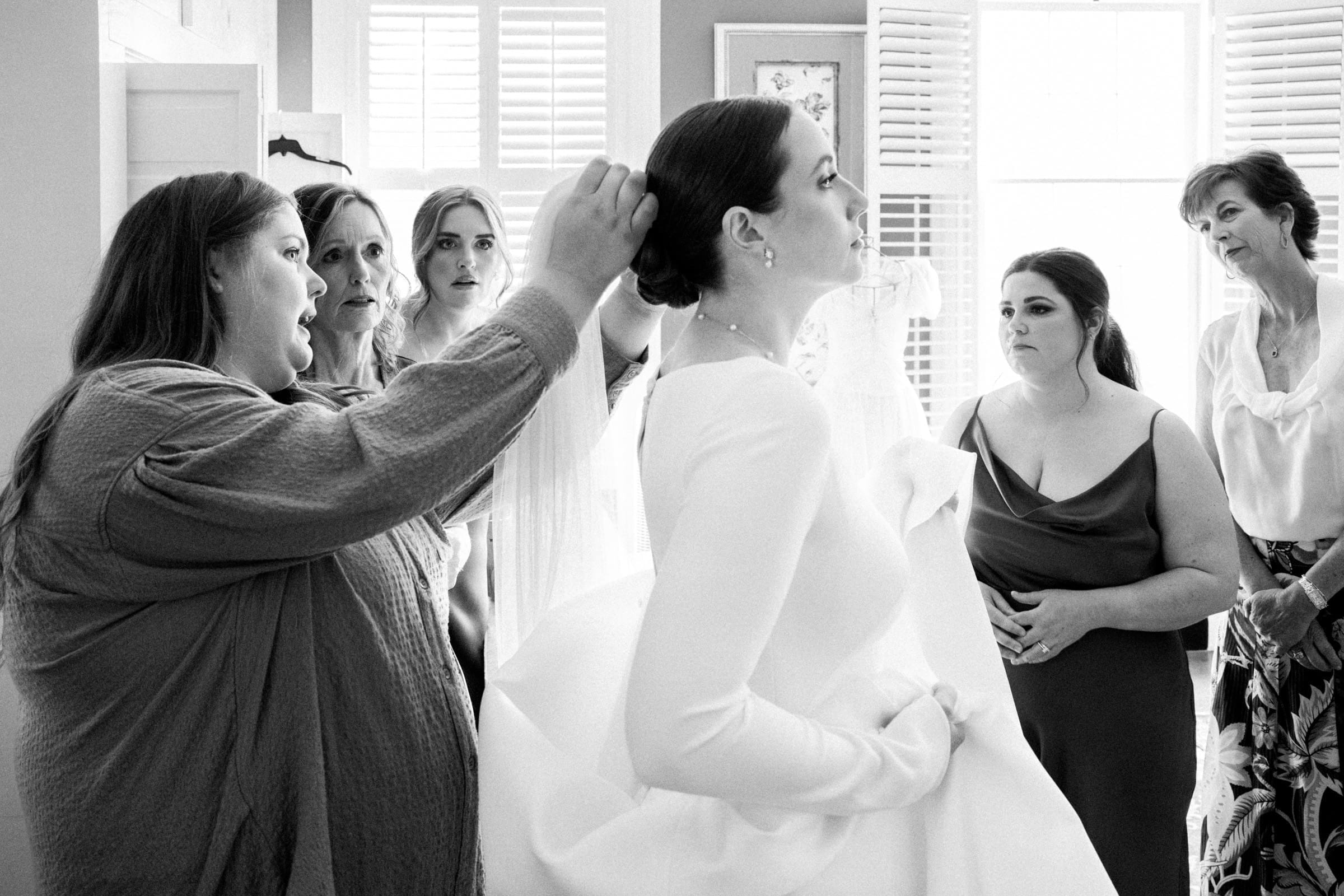 sutherland estate wedding - stephanie & hunter A black and white photo captures a Sutherland Estate wedding preparation scene. The bride, in her white dress, is surrounded by women assisting her. One adjusts the bride's hair while others offer support and chat in the background as light streams in from a window.