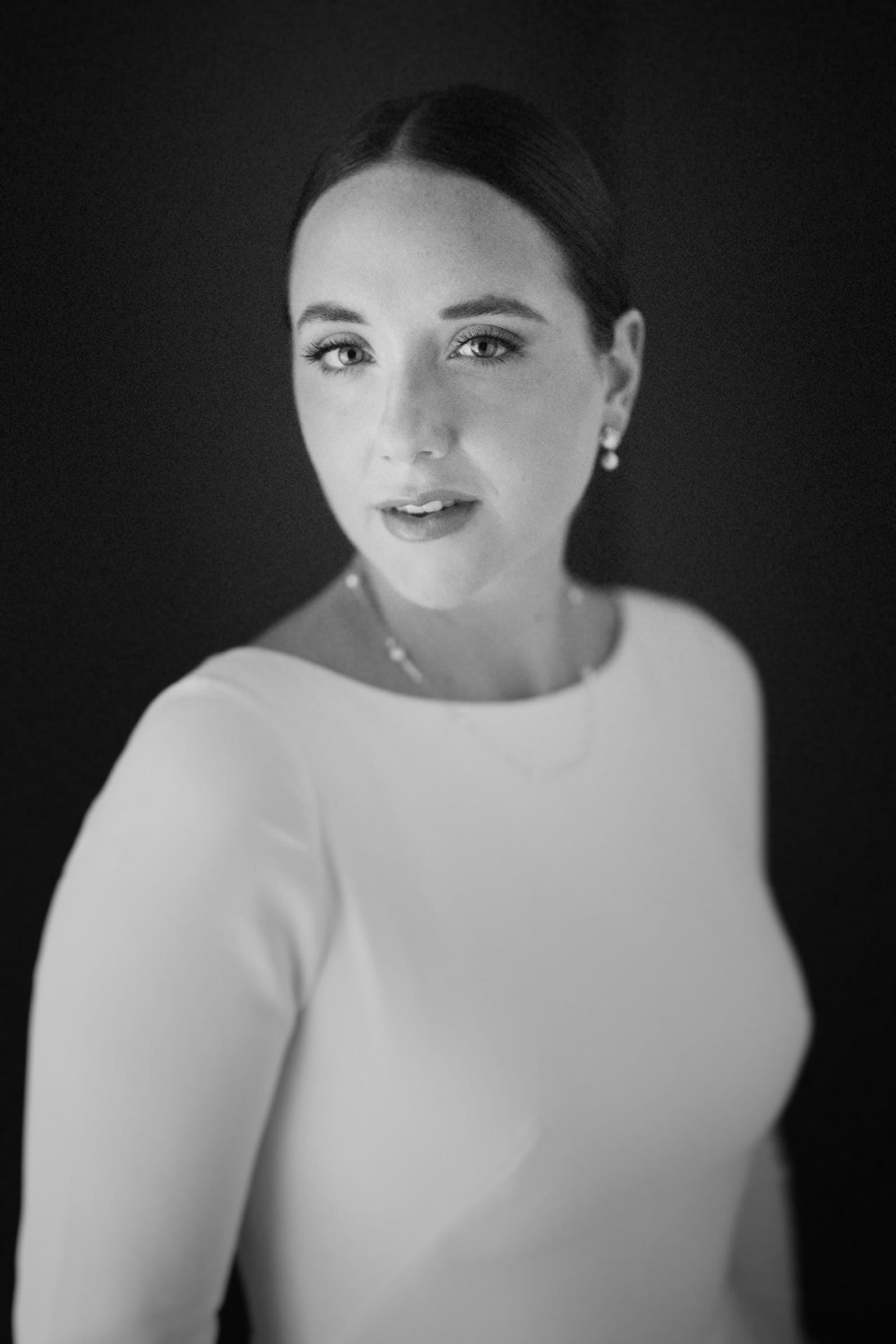 sutherland estate wedding - stephanie & hunter Black and white portrait of a person with dark hair in a low bun, wearing a white top and earrings. They are looking towards the camera with a neutral expression against a dark background, reminiscent of classic elegance found at a Sutherland Estate wedding.