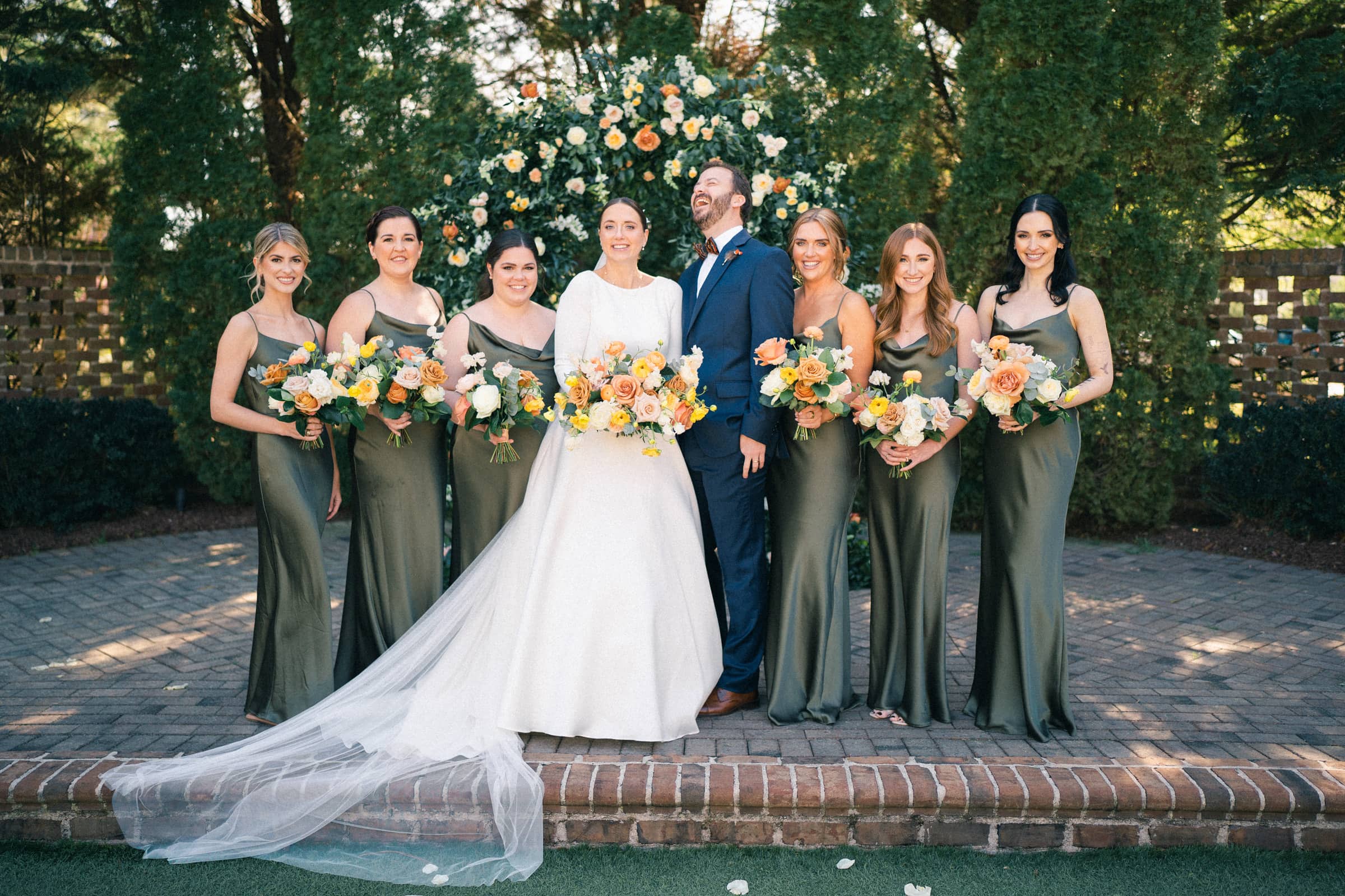 sutherland estate wedding - stephanie & hunter At their Sutherland Estate wedding, a bride and groom stand happily together outdoors in front of a floral arch. The bride is wearing a white dress, and the groom is in a blue suit. They are surrounded by six bridesmaids in matching green dresses, each holding bouquets.