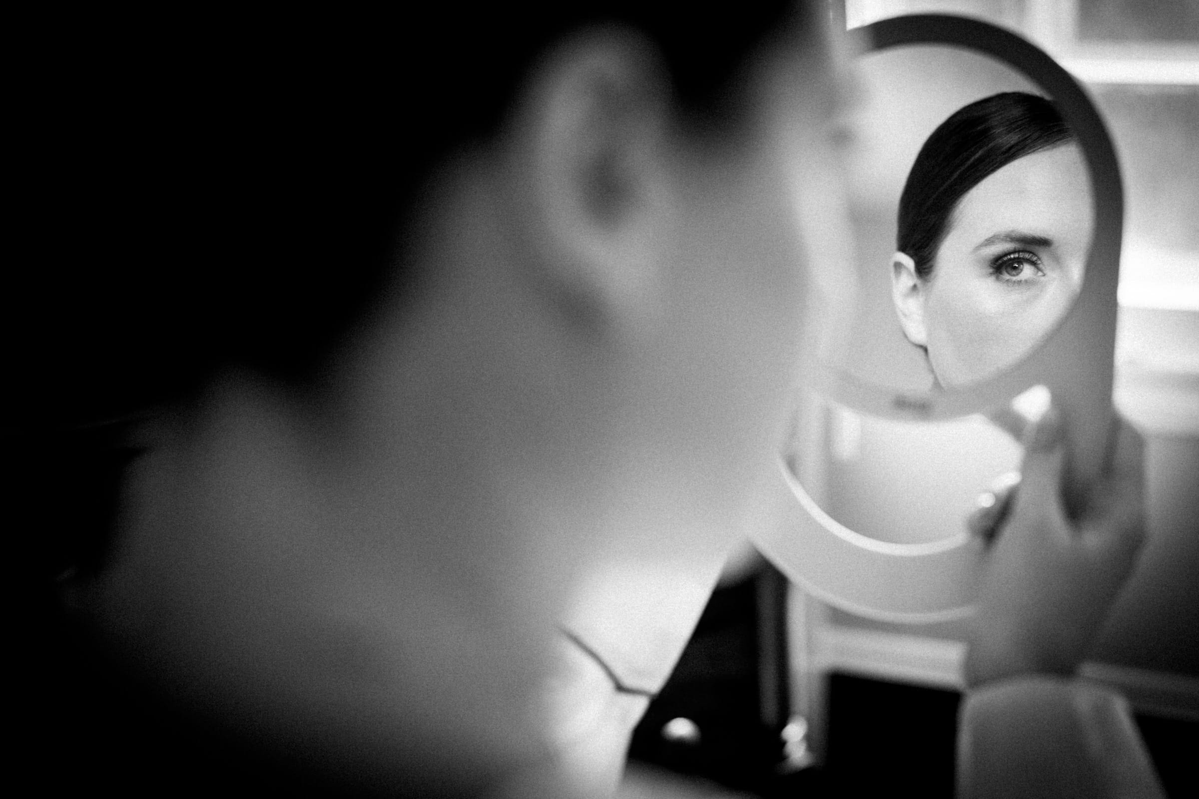 sutherland estate wedding - stephanie & hunter A black-and-white photo of a person at a Sutherland Estate wedding captures them holding a mirror close to their face. The mirror reflects one of their eyes, while the rest of the image is softly blurred, focusing attention on this intimate reflection amidst the celebration.