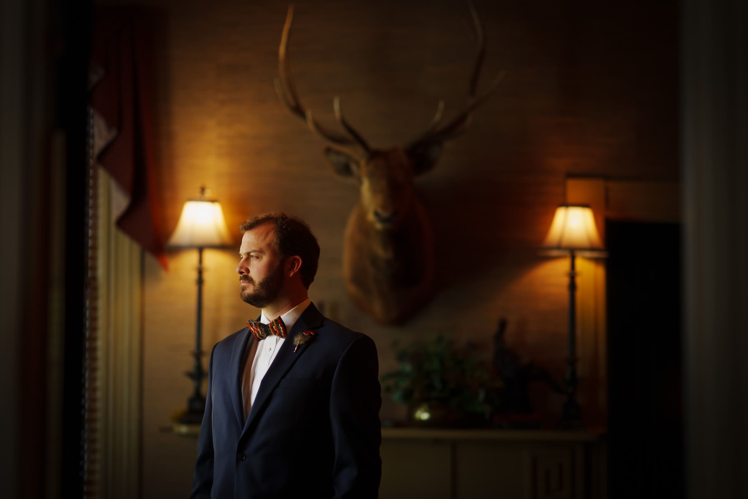 sutherland estate wedding - stephanie & hunter A man in a suit and bow tie stands in a dimly lit room, capturing the charm of a Sutherland Estate wedding. He looks to the side, with a mounted deer head and two lit lamps behind him, enhancing the room's warm, vintage atmosphere.