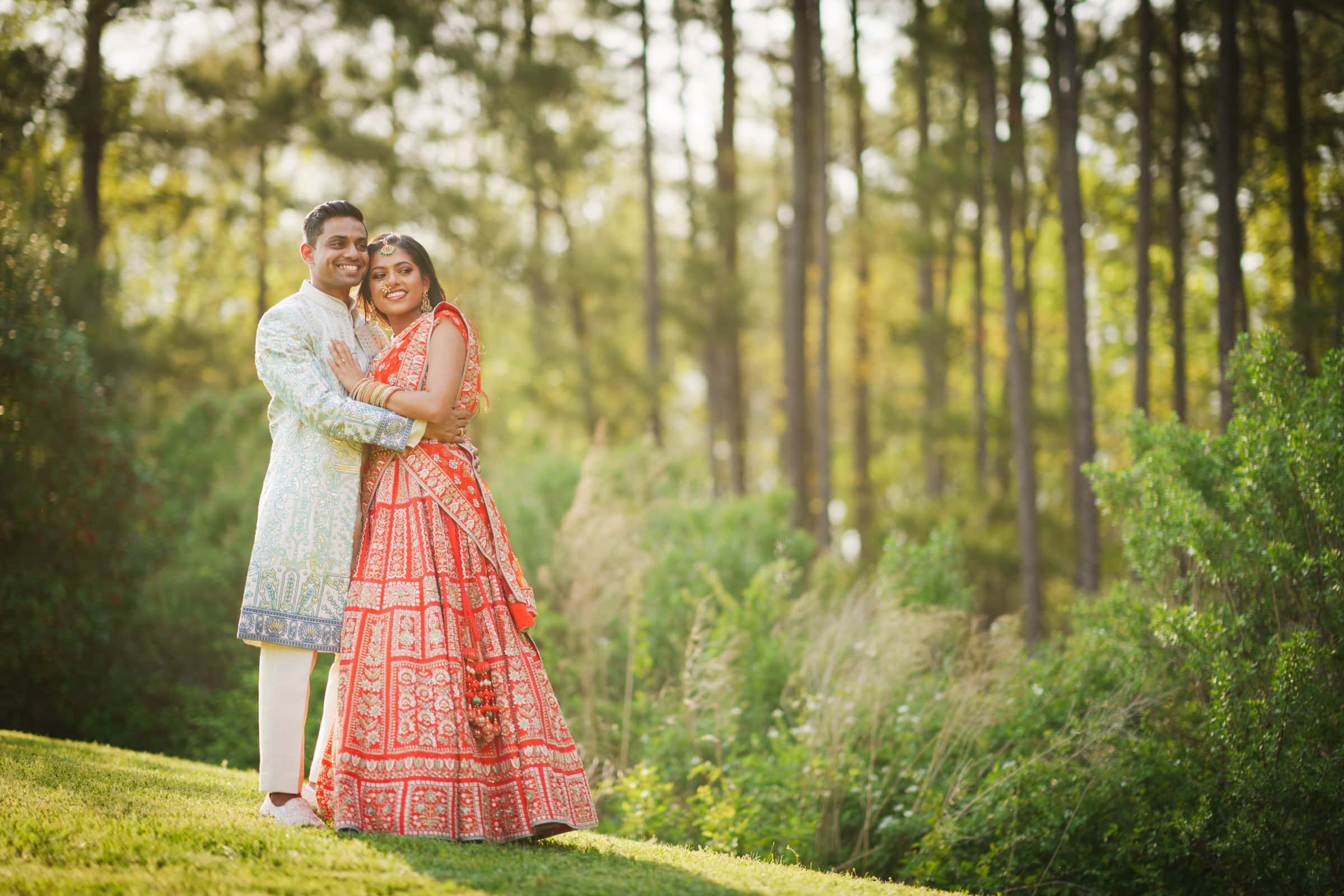 indian wedding photography - rithika & chanakya A couple in traditional Indian attire embraces happily on a grassy hill, capturing the essence of Indian wedding photography. The woman wears a vibrant red and gold lehenga, while the man dons a patterned sherwani. Tall trees and lush greenery frame this serene outdoor moment.