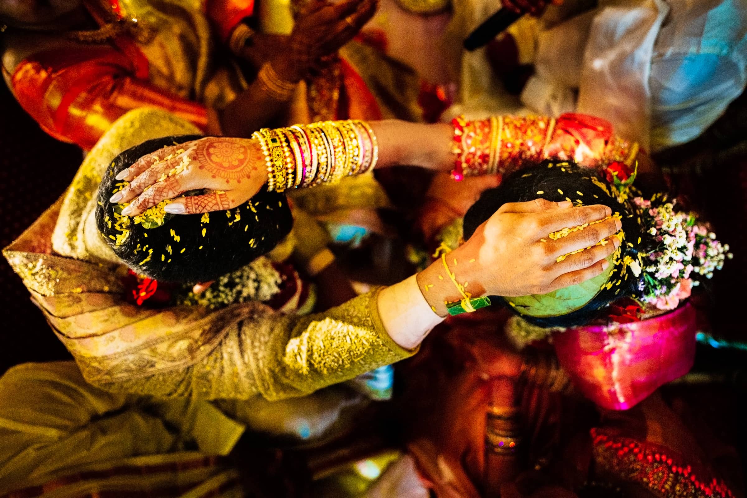A vibrant scene captured through Indian wedding photography showcases a bride and groom seated closely, adorned in traditional attire. Their heads are bowed under colorful flower petals, and intricate henna designs grace the bride's hand.