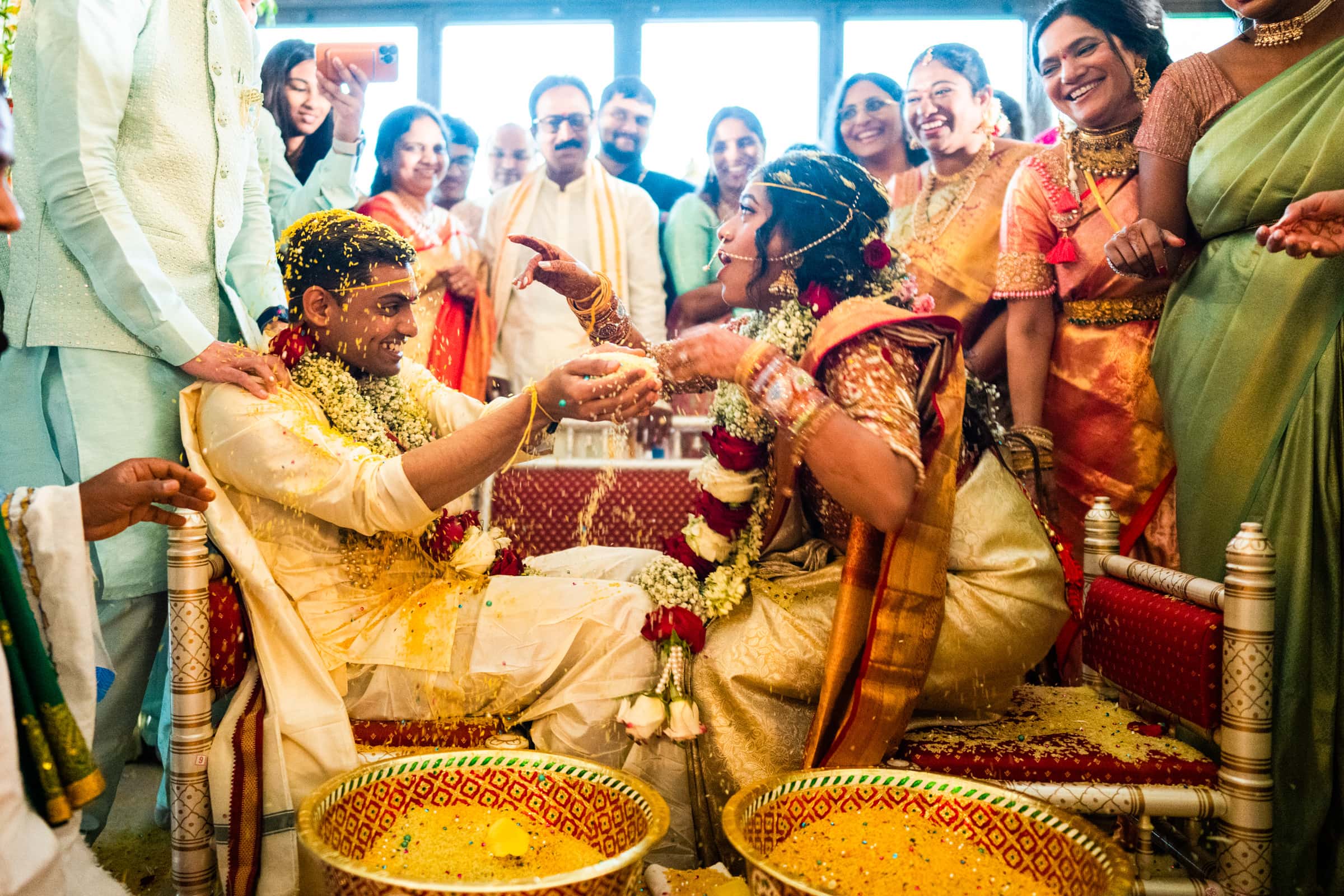 indian wedding photography - rithika & chanakya A couple dressed in traditional Indian attire participates in a joyful wedding ceremony, captured beautifully in Indian wedding photography. They are surrounded by smiling family and friends, with yellow rice scattered around. The atmosphere is festive and celebratory.