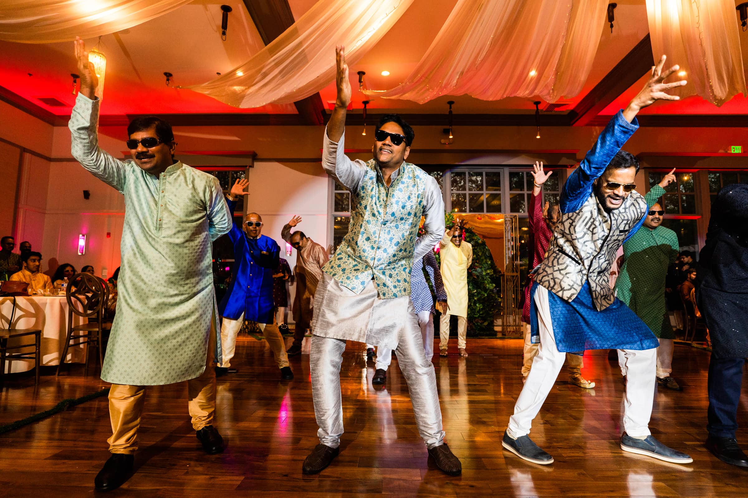 indian wedding photography - rithika & chanakya A group of men wearing colorful traditional attire are joyfully dancing with raised arms at a lively indoor event, captured in stunning Indian wedding photography. The scene is vibrant, with warm lighting and guests seated in the background.