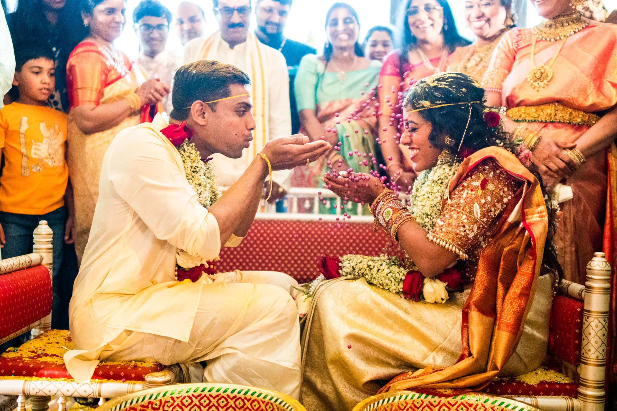 indian wedding photography - rithika & chanakya A bride and groom in traditional attire perform a wedding ritual, with marigold garlands and guests surrounding them. Captured by skilled Indian wedding photography, the groom gently blows petals towards the smiling bride, who is sitting opposite him.