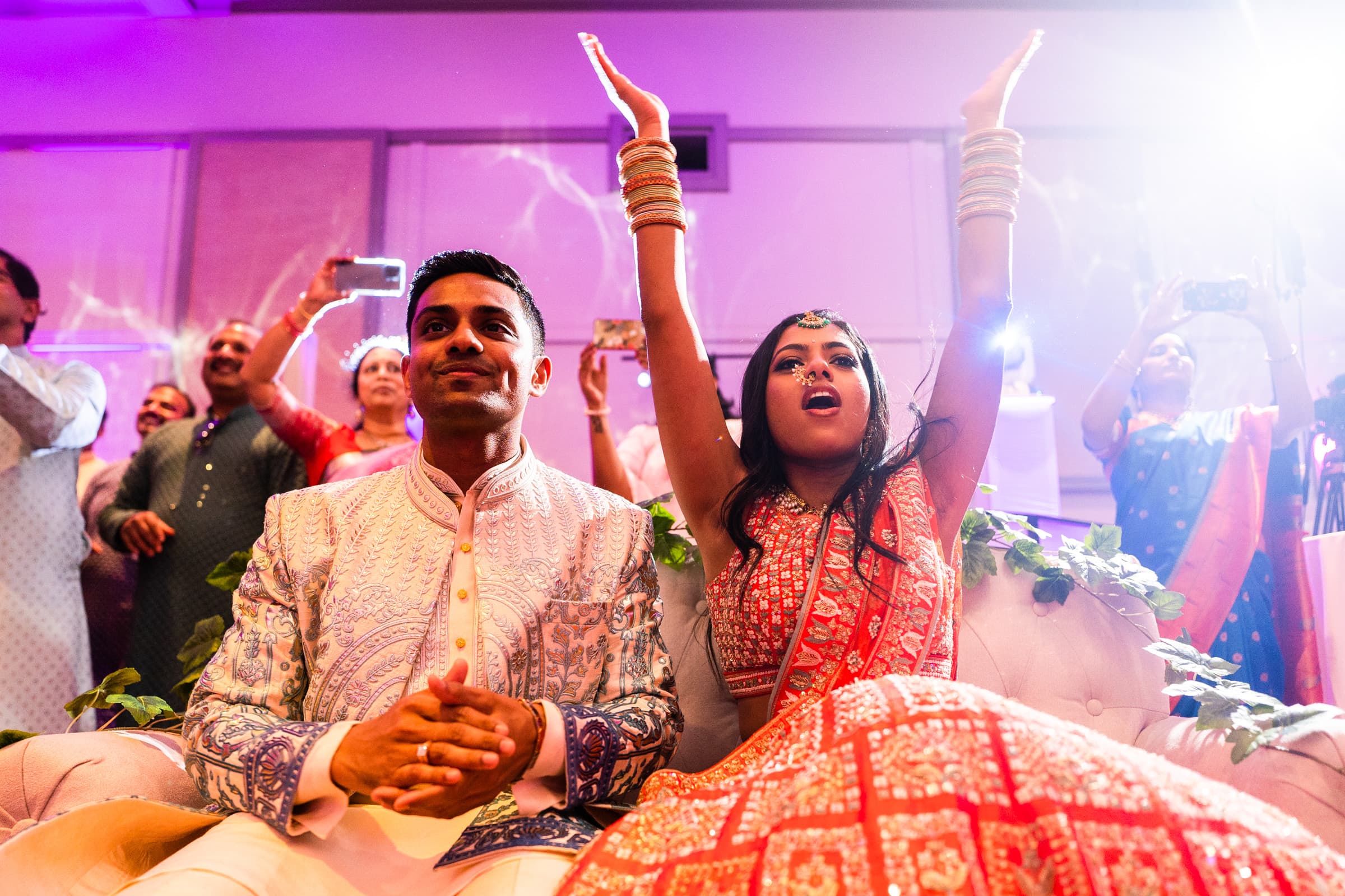 indian wedding photography - rithika & chanakya A couple in traditional attire sits on a couch at a lively event, perfectly capturing the essence of Indian wedding photography. The man wears a light blue embroidered sherwani, while the woman in a red lehenga raises her hands joyfully as people in the background snap photos.
