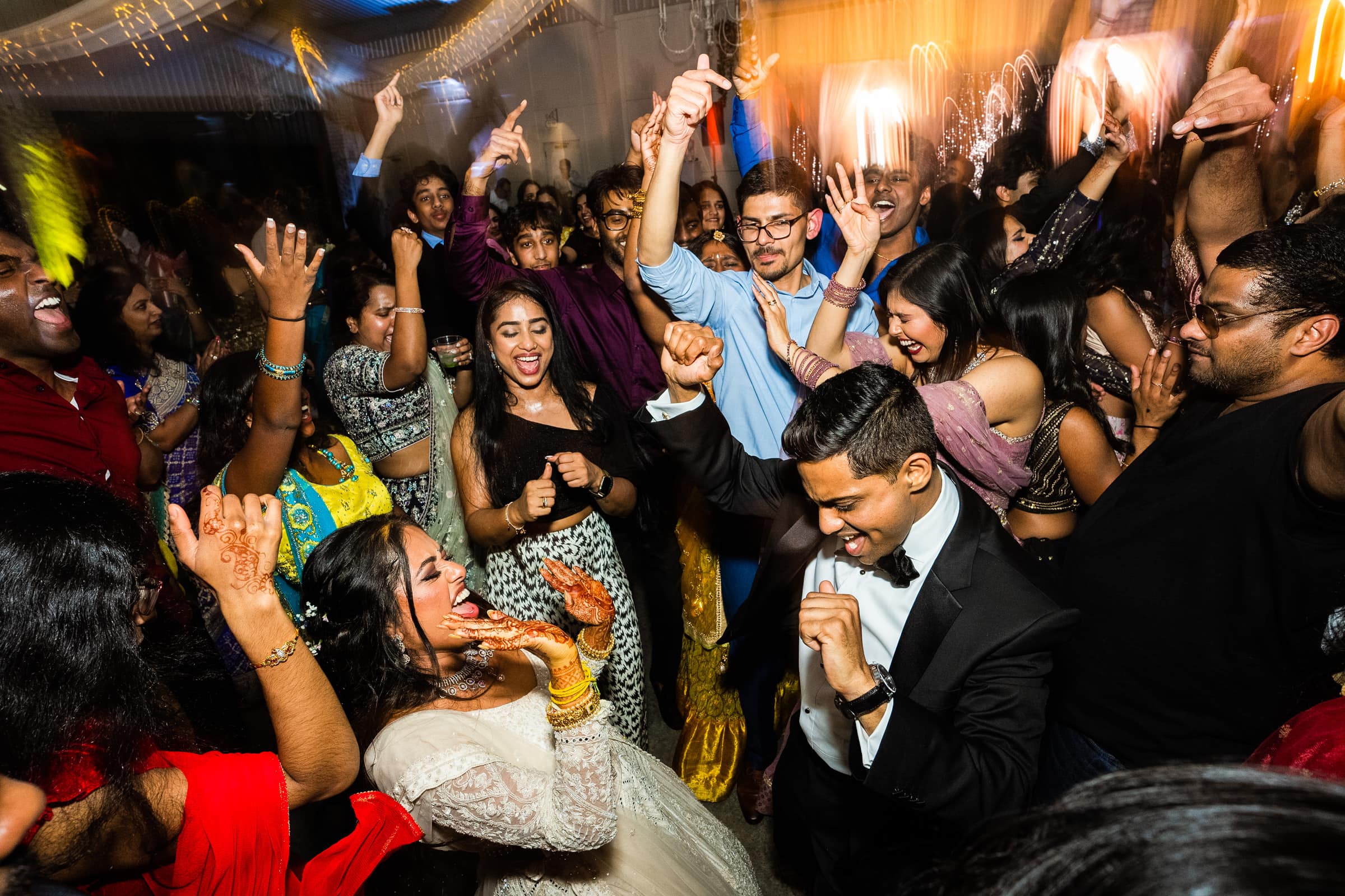 indian wedding photography - rithika & chanakya A lively Indian wedding photography scene captures the vibrant dance floor at a reception. Guests in colorful attire are joyfully dancing and celebrating. A woman in a white dress and a man in a suit are at the center, surrounded by smiling people with hands raised.