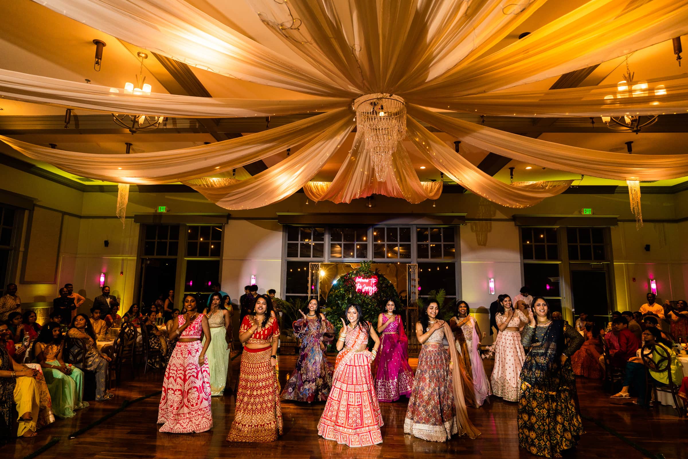 indian wedding photography - rithika & chanakya A group of women in colorful traditional dresses perform a choreographed dance at an indoor event, capturing moments perfect for Indian wedding photography. The venue's large drapes and elegant lighting add to the festive atmosphere, as an audience watches in awe from the background.