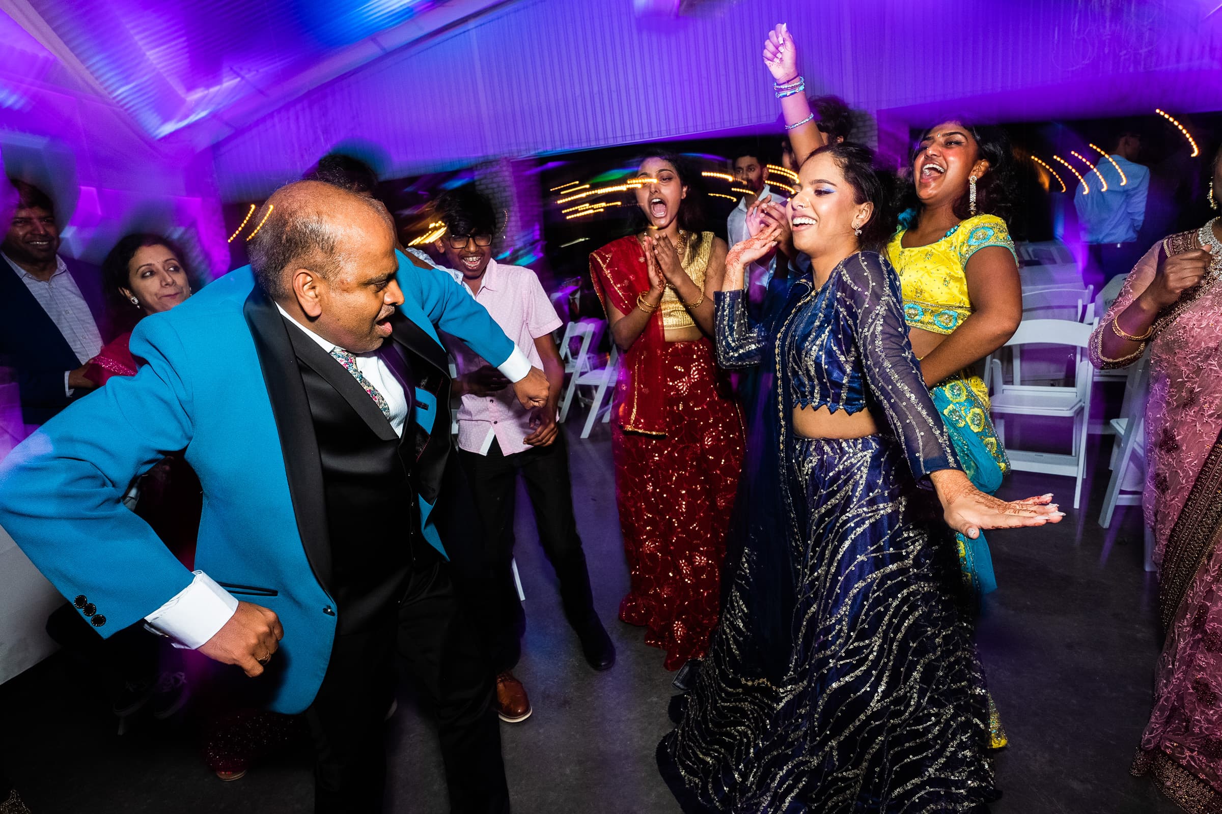 indian wedding photography - rithika & chanakya A group of people in colorful traditional attire are dancing energetically at what feels like an Indian wedding celebration. The scene is lively with music, as a man in a blue suit and a woman in a blue dress lead the dance under vibrant purple lighting, perfectly captured through photography.