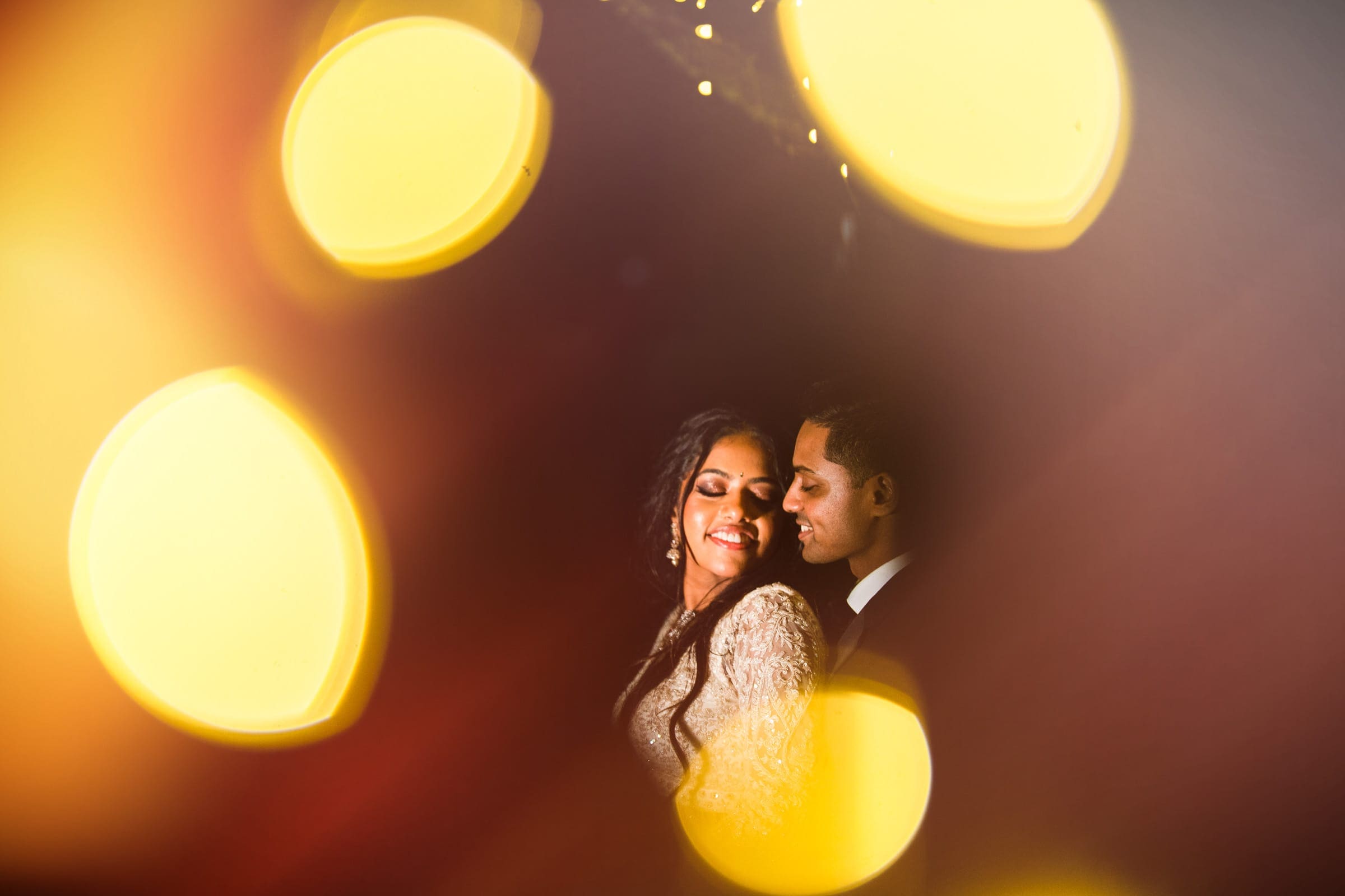indian wedding photography - rithika & chanakya A couple embraces in soft focus, surrounded by warm bokeh lights, capturing the essence of Indian wedding photography. The woman smiles gently with her eyes closed, while the man stands closely behind her, creating a romantic atmosphere.