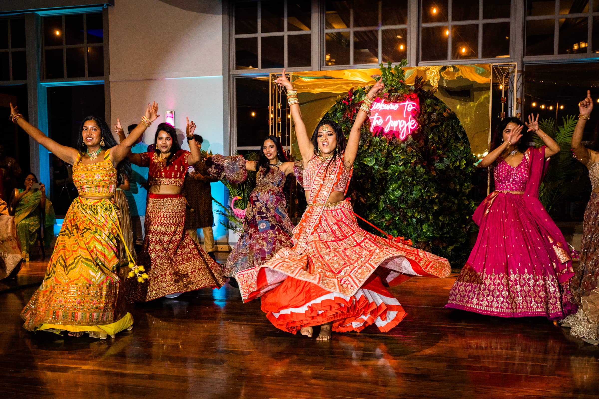 indian wedding photography - rithika & chanakya A lively group of women in colorful traditional outfits perform a dance at an Indian wedding. They are in motion, with flowing skirts twirling. A neon sign in the background and bright lights add to the vibrant atmosphere, perfectly captured by wedding photography.