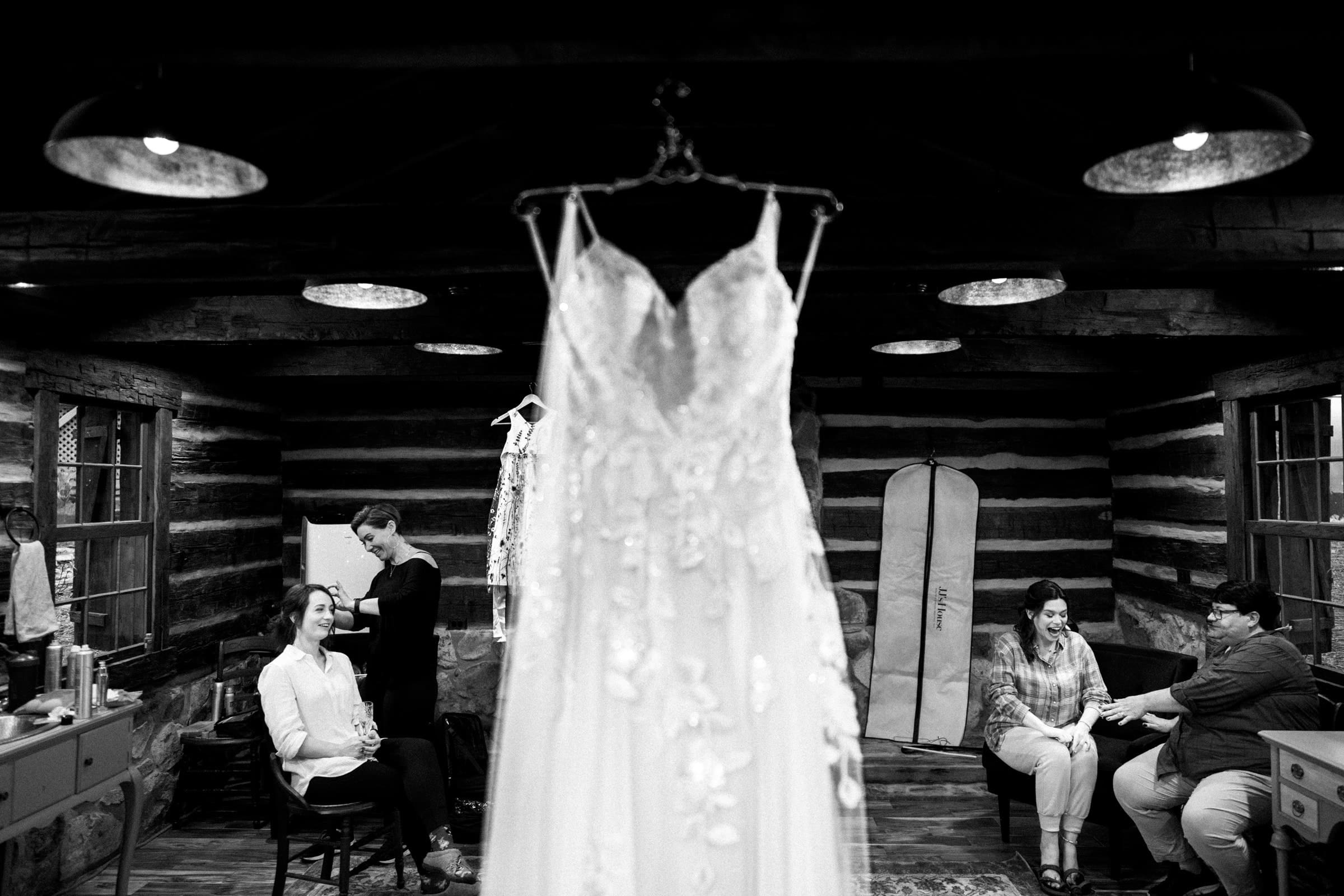 union grove farm wedding photography - kaylyn & jeremy A wedding dress hangs in focus at the center of a rustic Union Grove Farm room. Three people sit in the background; one is getting their hair styled while two others are engaged in conversation, capturing a serene moment of togetherness through the lens of wedding photography.