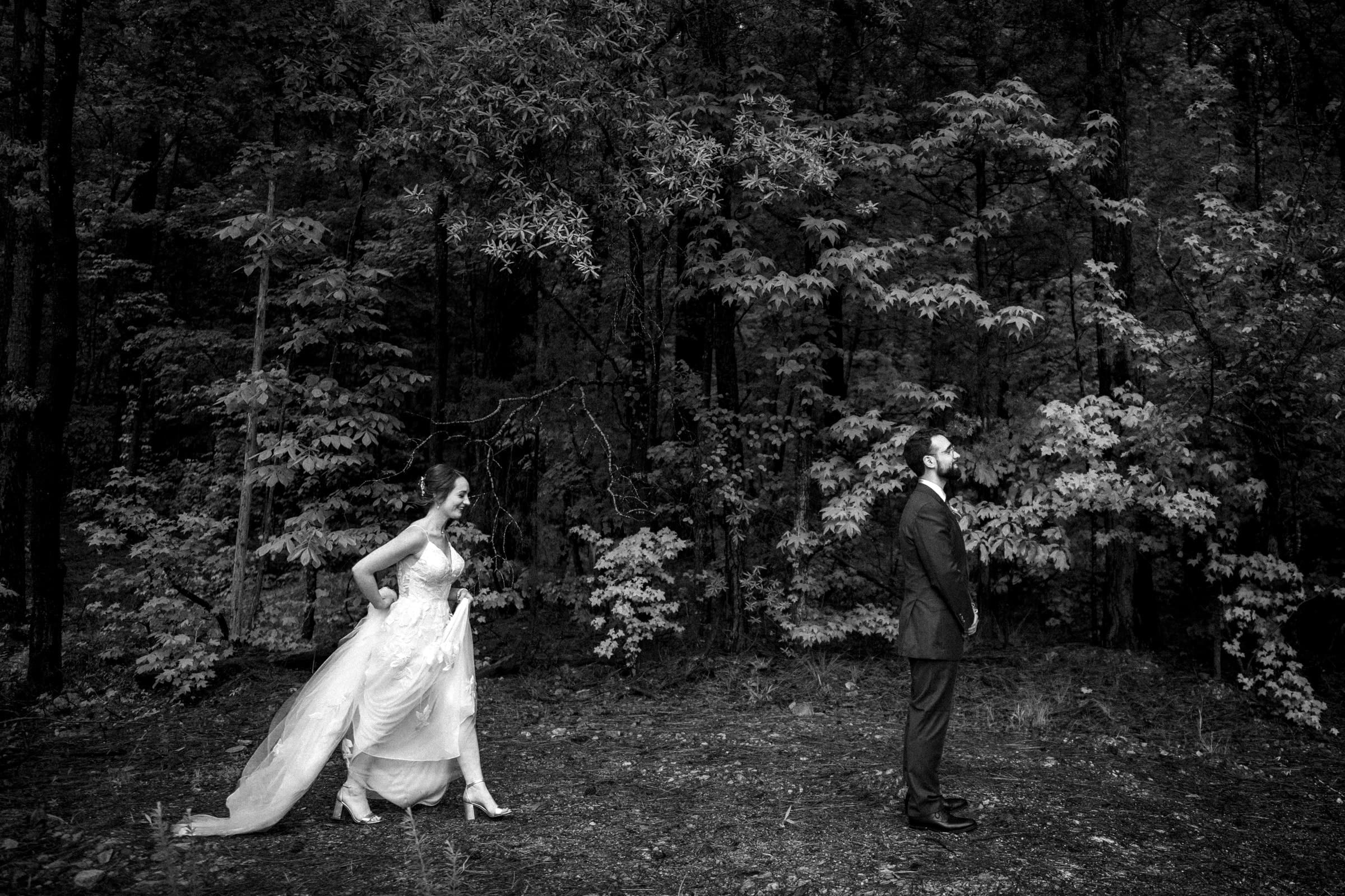 union grove farm wedding photography - kaylyn & jeremy A black and white photo captures a bride approaching her groom from behind in a lush, forest setting. Her flowing dress and heels contrast subtly with his suit amidst the tall trees' dense foliage, offering a timeless moment of union reminiscent of classic grove farm wedding photography.