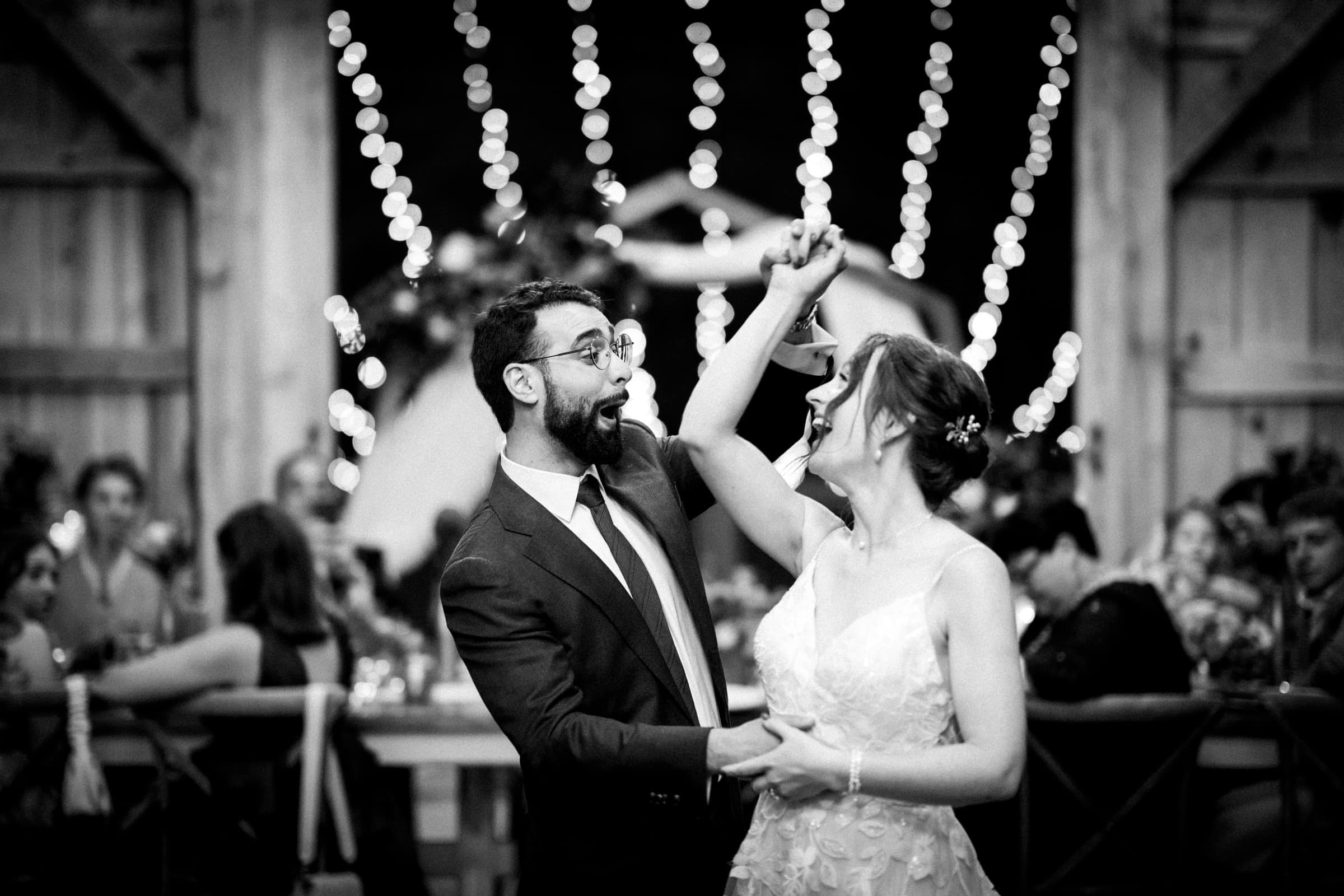 union grove farm wedding photography - kaylyn & jeremy A couple dances joyfully at a Union Grove Farm wedding reception. The man twirls the woman under a canopy of string lights, perfectly captured in photography. Guests can be seen seated in the background, adding to the festive atmosphere.