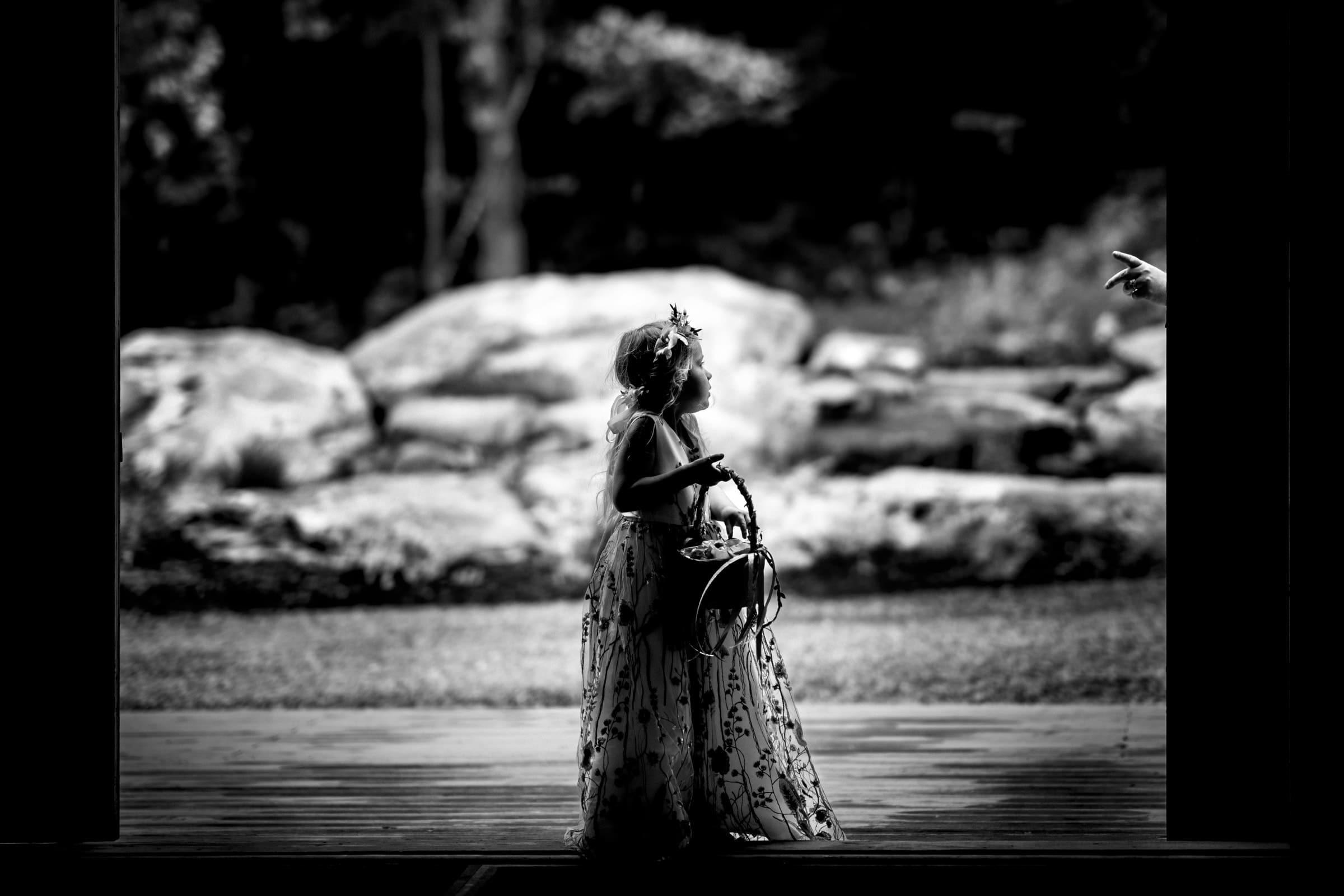 union grove farm wedding photography - kaylyn & jeremy A black and white photo captures a young girl in a floral dress, holding a basket, standing indoors near a doorway. Her gaze is fixed on an extended hand, with the rocky terrain of Union Grove Farm framing the background, adding a rustic charm to this wedding photography scene.