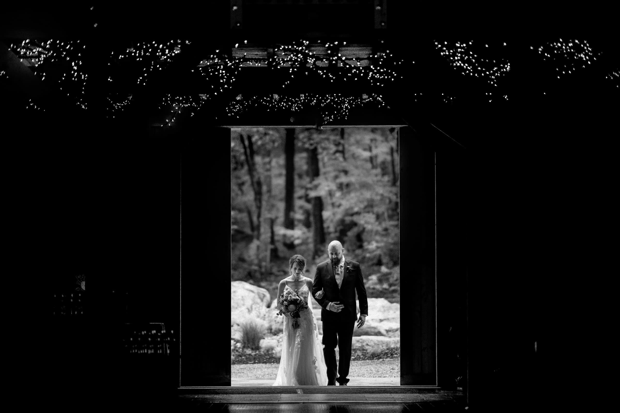 union grove farm wedding photography - kaylyn & jeremy A black-and-white photo captures a bride and an older man walking down an aisle, framed by a doorway with twinkling lights above. This enchanting scene at Union Grove Farm leads to a forested outdoor setting, where the bride holds a bouquet, preserving the magic of wedding photography.