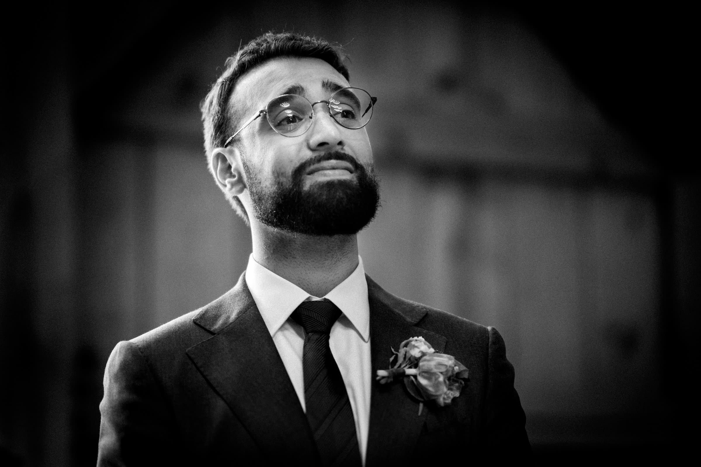 union grove farm wedding photography - kaylyn & jeremy A man with glasses, a beard, and a formal suit adorned with a floral boutonniere gazes upward. Captured in black and white by Union Grove’s finest wedding photography, the image reveals his thoughtful expression against a softly blurred background.