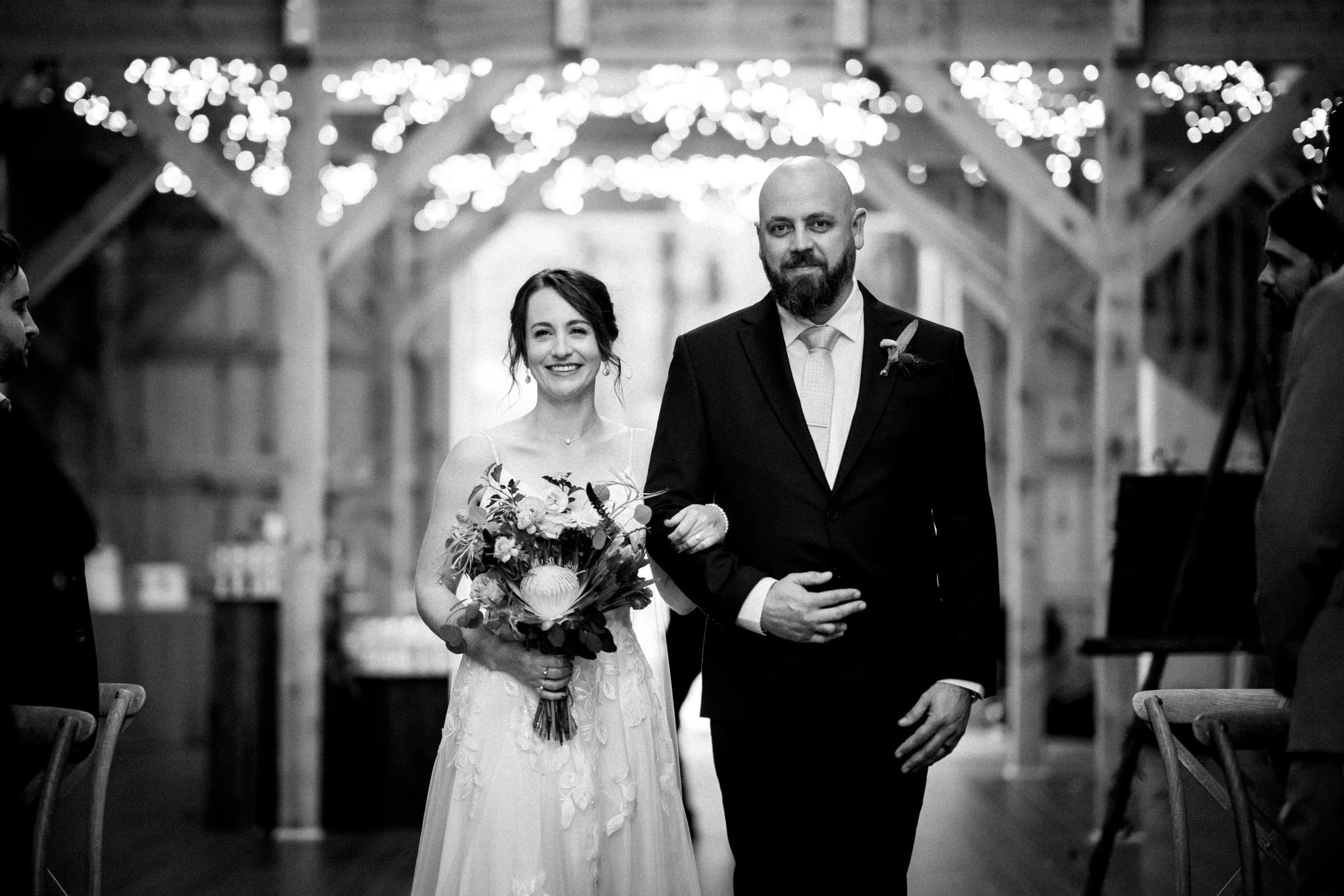 union grove farm wedding photography - kaylyn & jeremy A bride and groom walk down the aisle at Union Grove Farm, embraced by a warmly lit, rustic setting. The bride holds a bouquet and smiles, while the groom is beside her. Twinkling lights create a cozy atmosphere in the background, captured beautifully in black and white wedding photography.