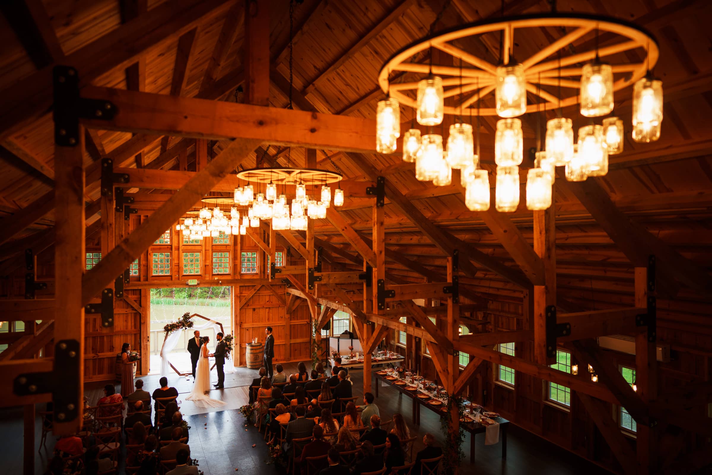 union grove farm wedding photography - kaylyn & jeremy A warm, cozy union ceremony unfolds in a rustic barn. Guests are seated, facing the couple at the altar. Hanging wagon wheel chandeliers with glowing lights illuminate the wooden interior of Grove Farm, creating a romantic atmosphere perfect for capturing in wedding photography.