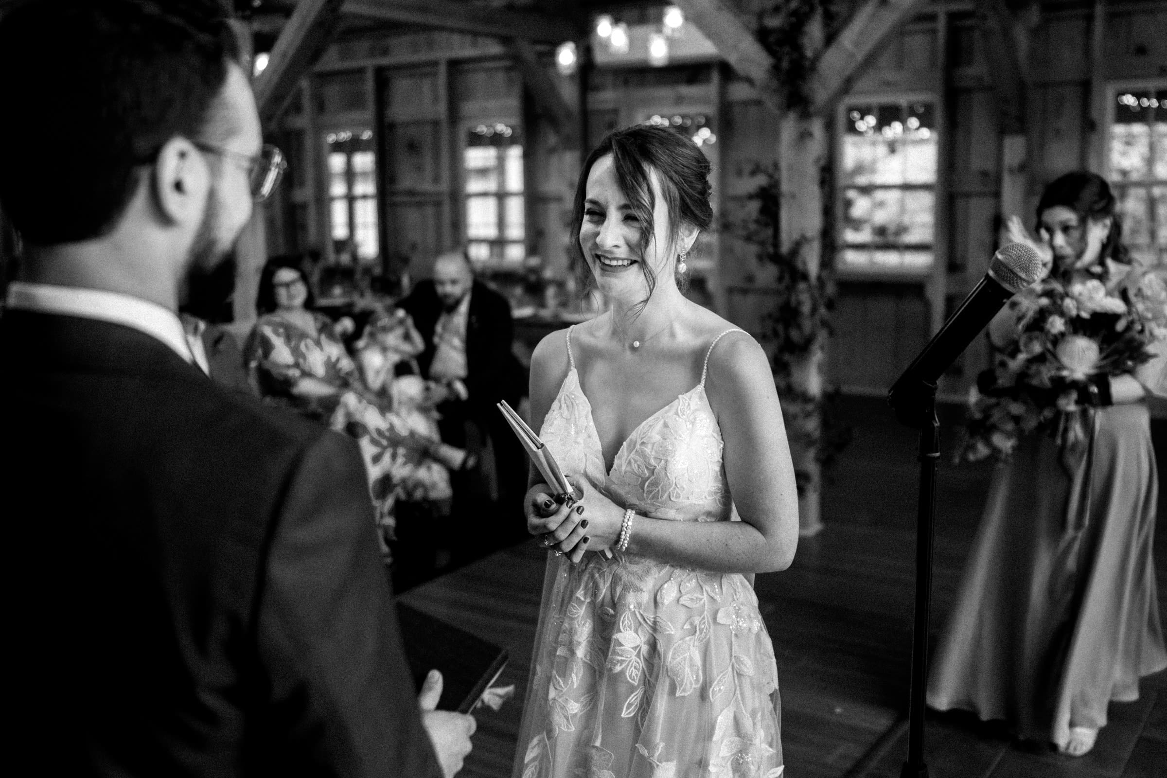 union grove farm wedding photography - kaylyn & jeremy A bride smiles warmly as she reads her vows to the groom during their intimate Union Grove Farm ceremony. Holding a notepad, with a bridesmaid in the background clutching a bouquet, the moment is beautifully captured by wedding photography amid an atmosphere of joy.