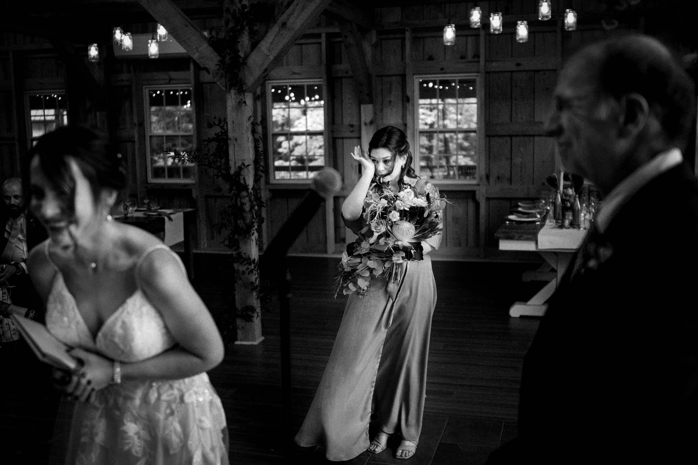 union grove farm wedding photography - kaylyn & jeremy In a rustic venue with wooden beams at Union Grove Farm, a woman in a sleeveless dress wipes a tear while holding a large bouquet. A bride and an older man are in the foreground, slightly blurred, capturing an emotional moment in wedding photography.