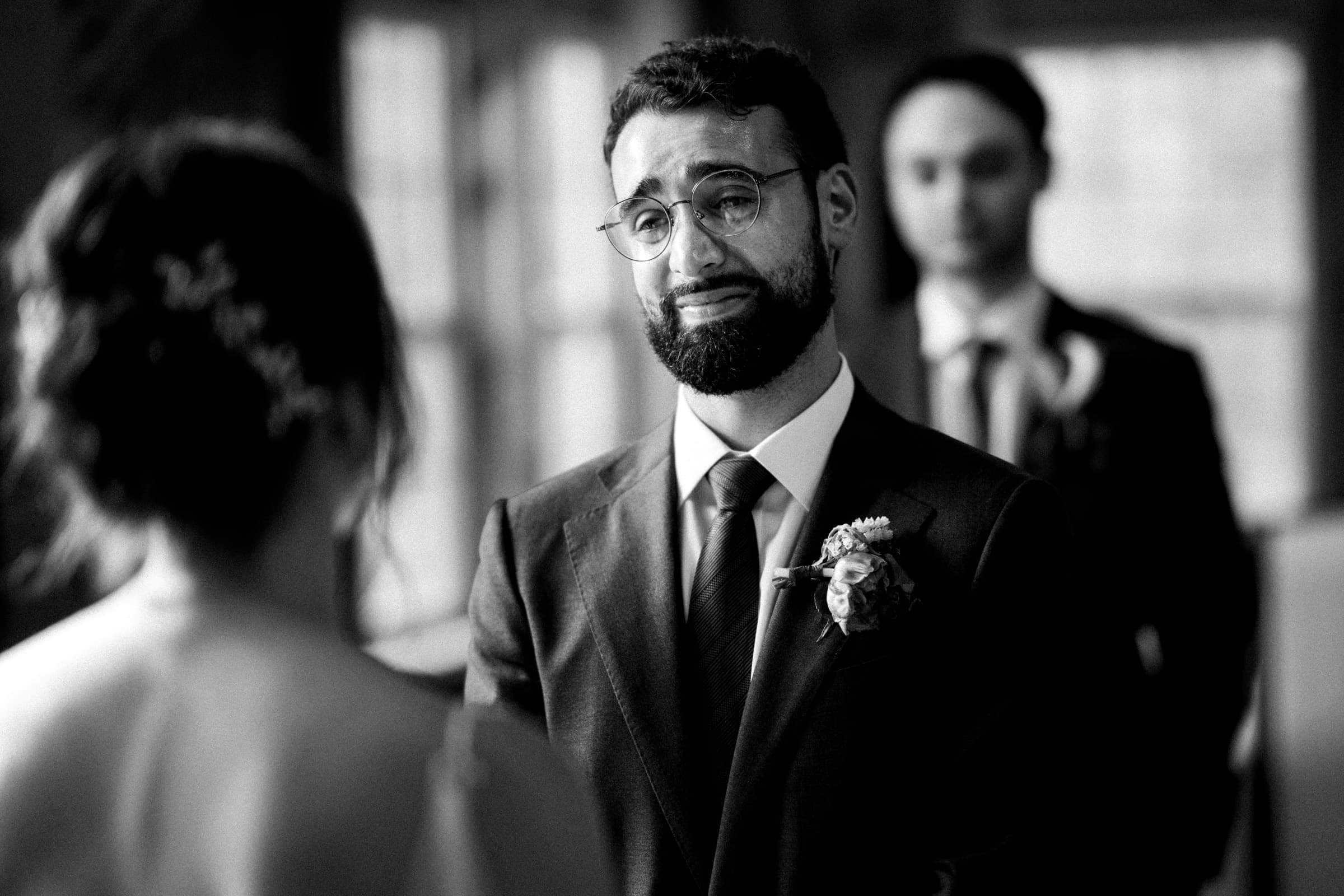 union grove farm wedding photography - kaylyn & jeremy A groom in a suit and glasses stands facing a bride during their Union Grove Farm ceremony, appearing emotional. A groomsman is blurred in the background. The indoor setting features soft lighting, captured beautifully in black and white wedding photography.