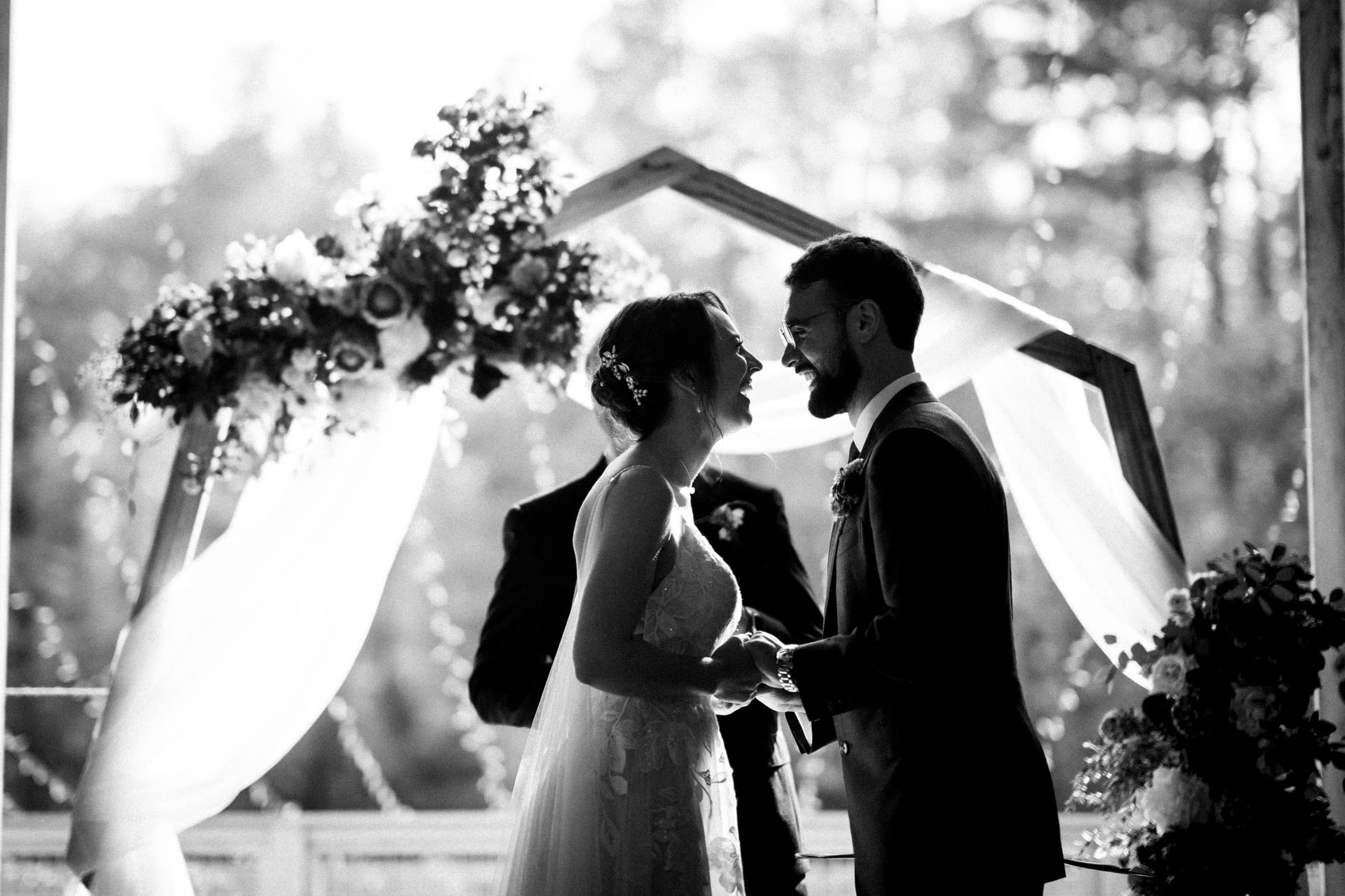 union grove farm wedding photography - kaylyn & jeremy A couple stands facing each other and smiling during their outdoor wedding ceremony at Union Grove Farm. They are under a wooden geometric arch adorned with flowers and draped fabric, creating a picturesque scene with blurred trees and soft natural light in the background.