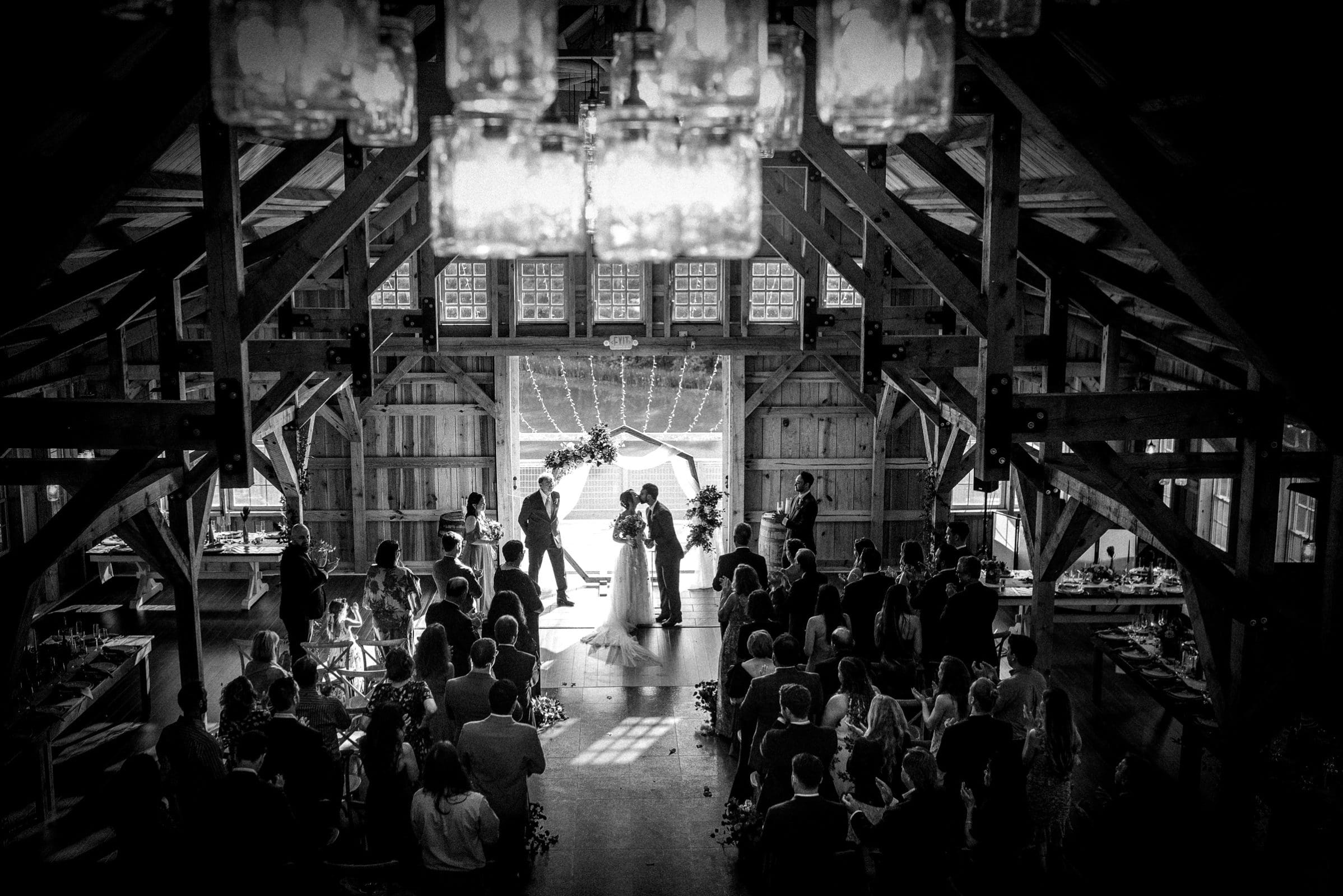 union grove farm wedding photography - kaylyn & jeremy A black and white photo captures a beautiful union at a rustic barn wedding. The couple shares a kiss at the altar while guests sit on either side. Sunlight streams through open barn doors at Grove Farm, illuminating the scene as hanging lights create a warm, intimate atmosphere.