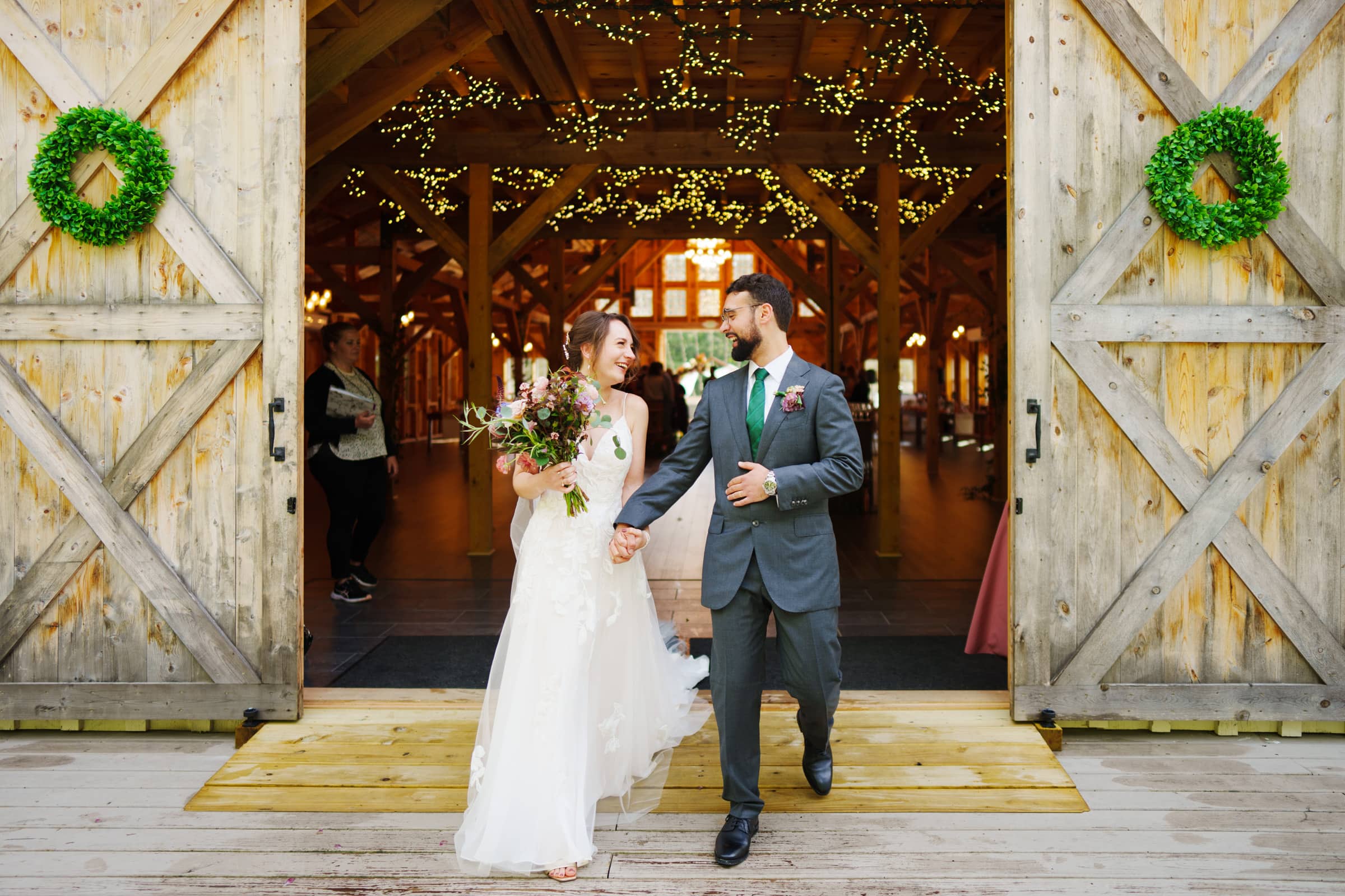 union grove farm wedding photography - kaylyn & jeremy A bride and groom joyfully exit a wooden barn at Union Grove Farm, decorated with wreaths and twinkling lights. The bride holds a bouquet, their smiles captured perfectly by wedding photography as they hold hands. The barn's open double doors reveal greenery inside.