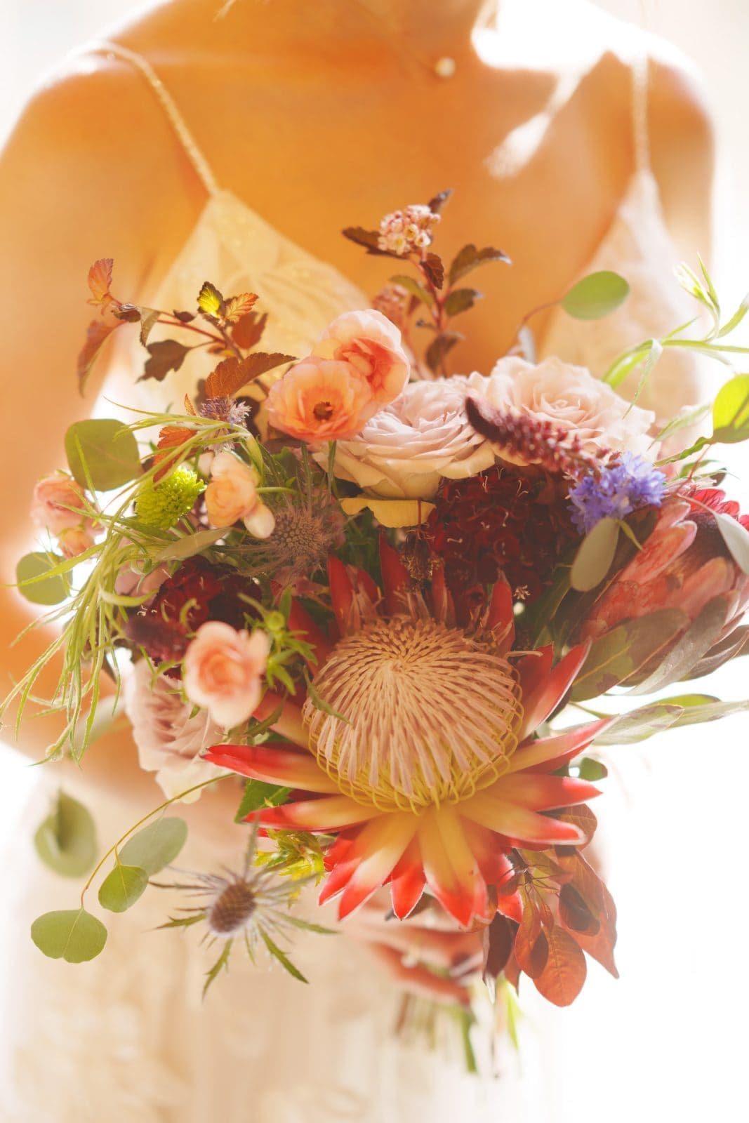 union grove farm wedding photography - kaylyn & jeremy A person in a white lace dress holds a vibrant bouquet of flowers, including pink roses, red blooms, purple thistle, and green leaves. Sunlight softly illuminates the bouquet at Union Grove Farm, highlighting its diverse textures and colors—perfect for wedding photography.