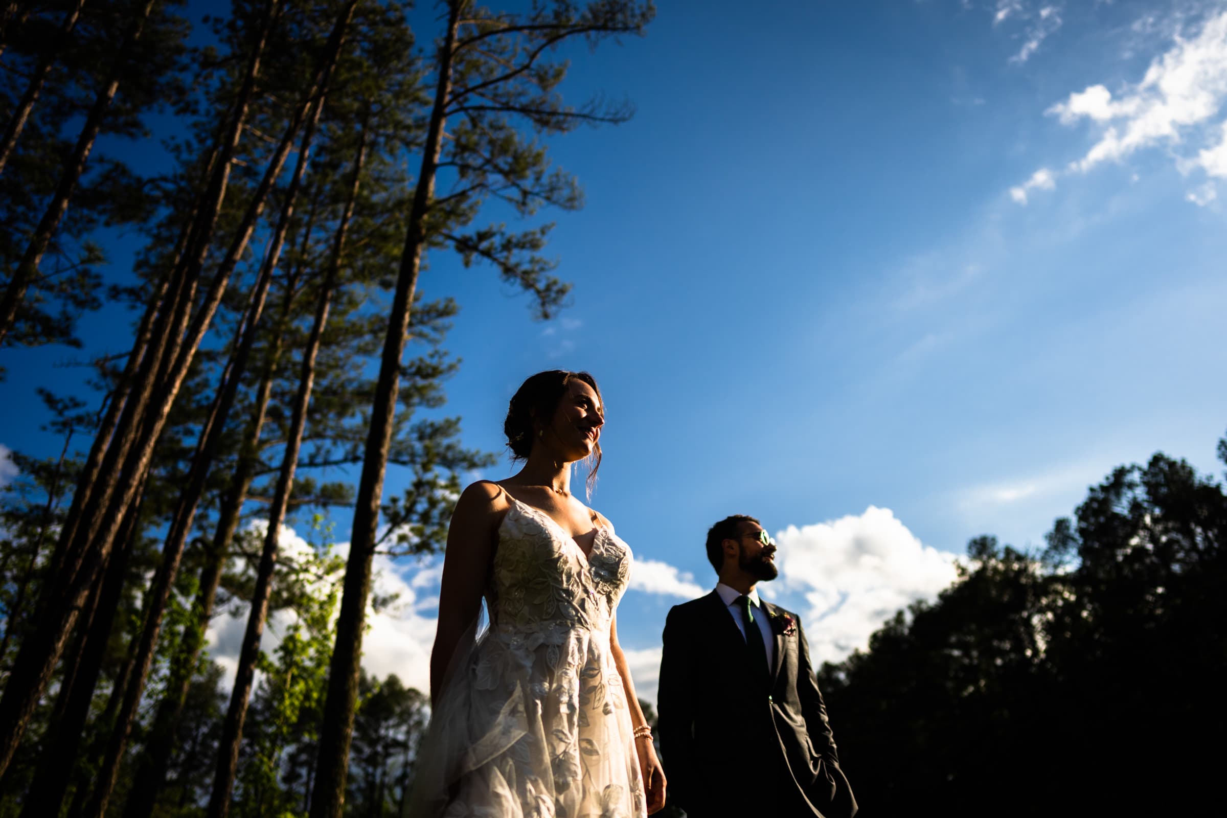 union grove farm wedding photography - kaylyn & jeremy A couple stands outdoors in formal attire at Union Grove Farm, with tall trees and a bright blue sky as the stunning backdrop. The woman is wearing a white dress, and the man is in a dark suit. Sunlight casts dramatic shadows, highlighting the scene's natural beauty—perfect for wedding photography.