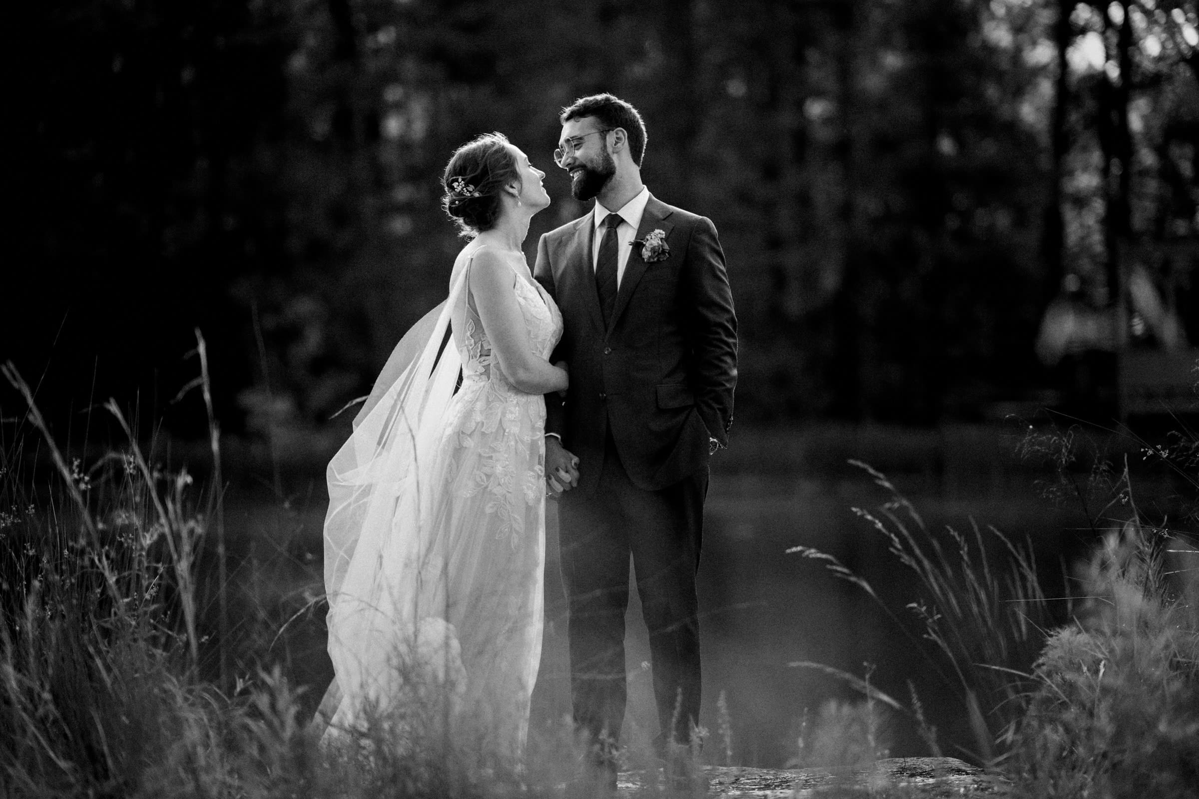 union grove farm wedding photography - kaylyn & jeremy A black and white photo captures a serene moment of a bride and groom standing hand in hand by a lake. The bride's flowing gown contrasts beautifully with the groom's suit adorned with a boutonniere. Amidst tall grass and trees, this union is beautifully immortalized by Grove Farm Wedding Photography.