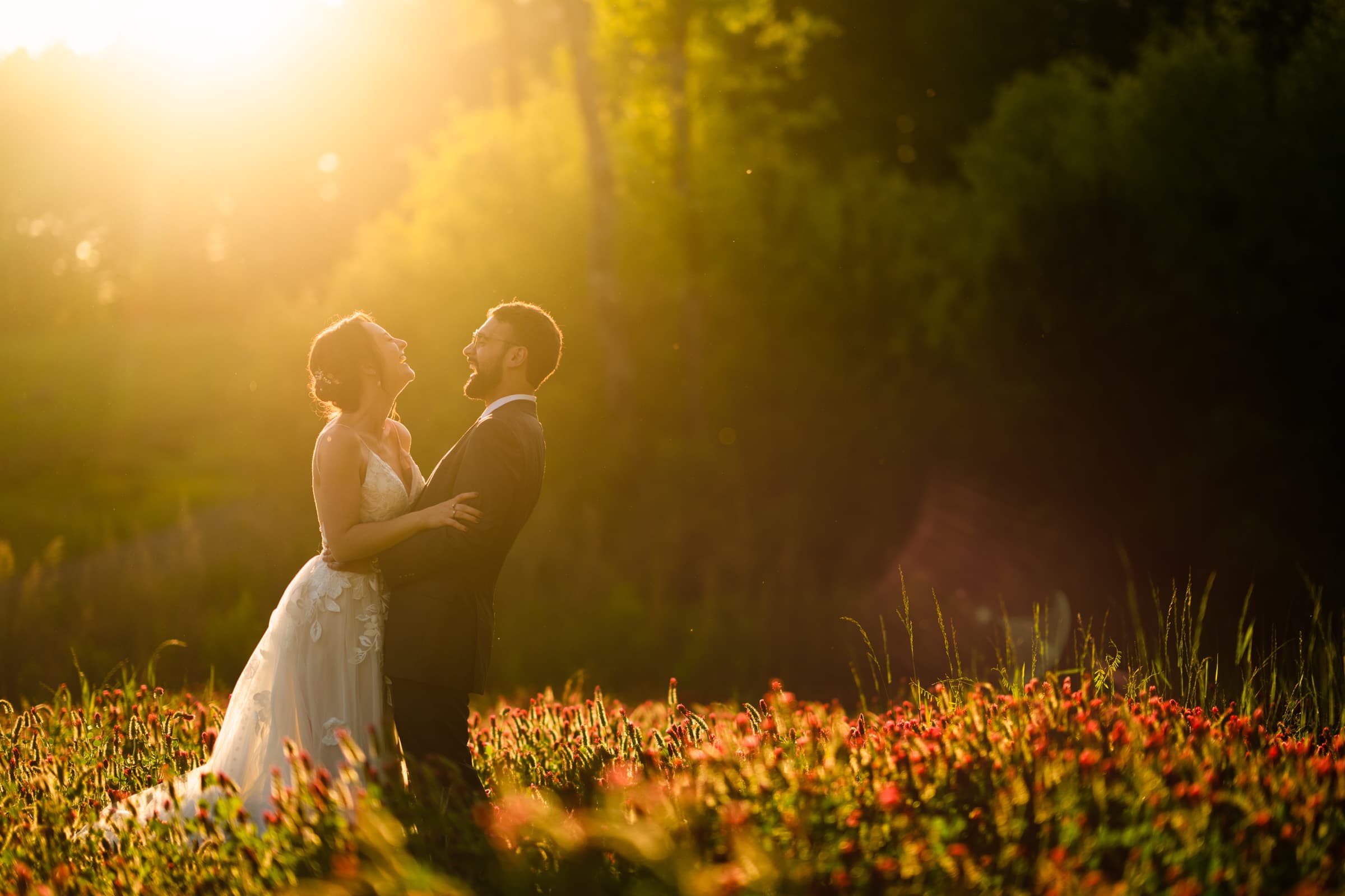 union grove farm wedding photography - kaylyn & jeremy A couple stands in a field of red and green wildflowers, embraced and smiling at each other. The warm, golden sunlight streams through the trees at Union Grove Farm, creating a romantic and serene atmosphere perfect for wedding photography.